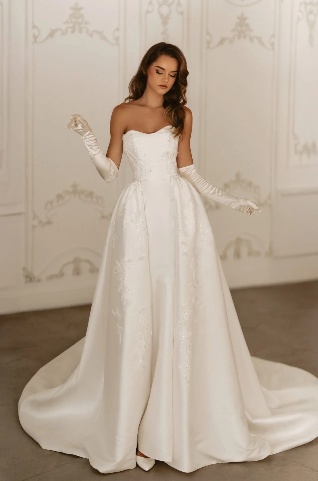 Young woman in a strapless white wedding gown with gloves, standing in an ornate room.