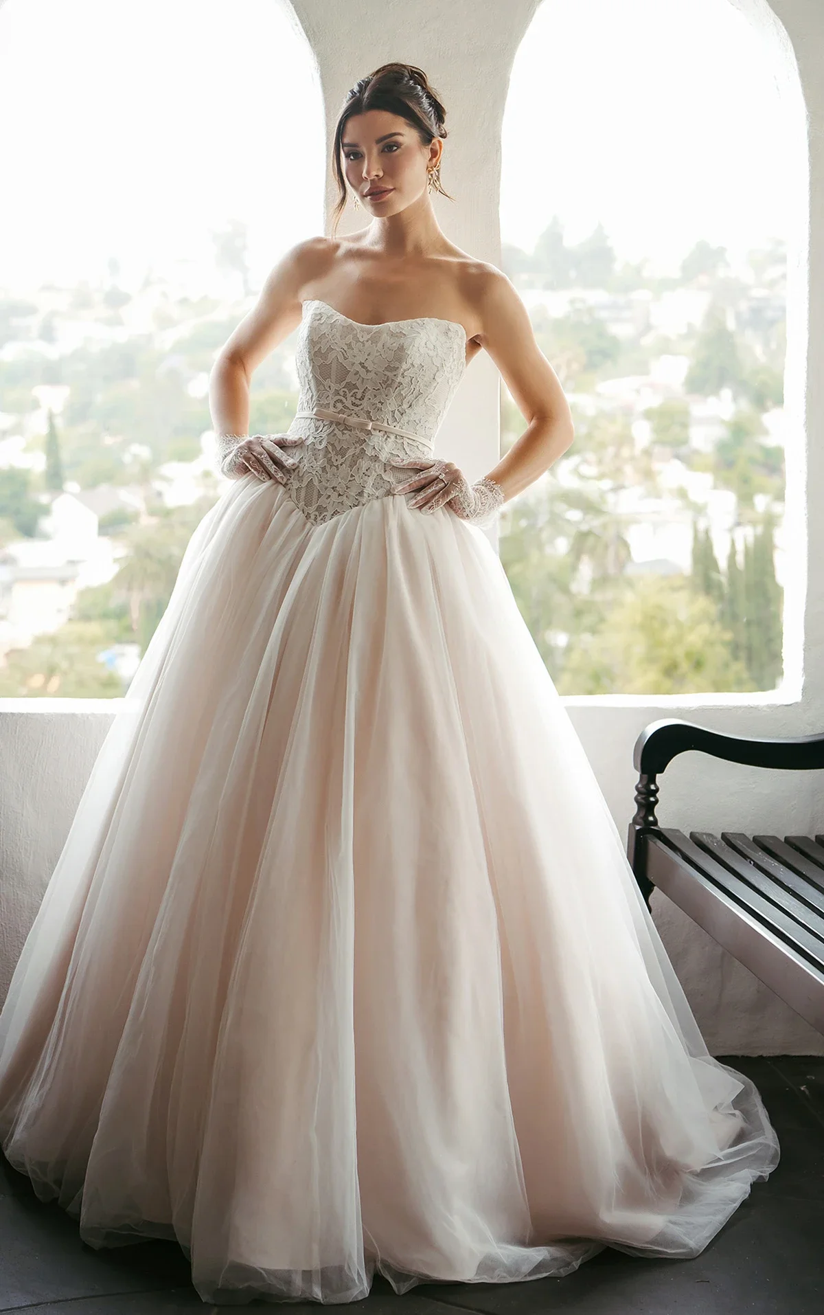 Woman in a strapless wedding gown with lace bodice and full tulle skirt, standing by a large window with an outdoor view, wearing lace gloves and earrings.