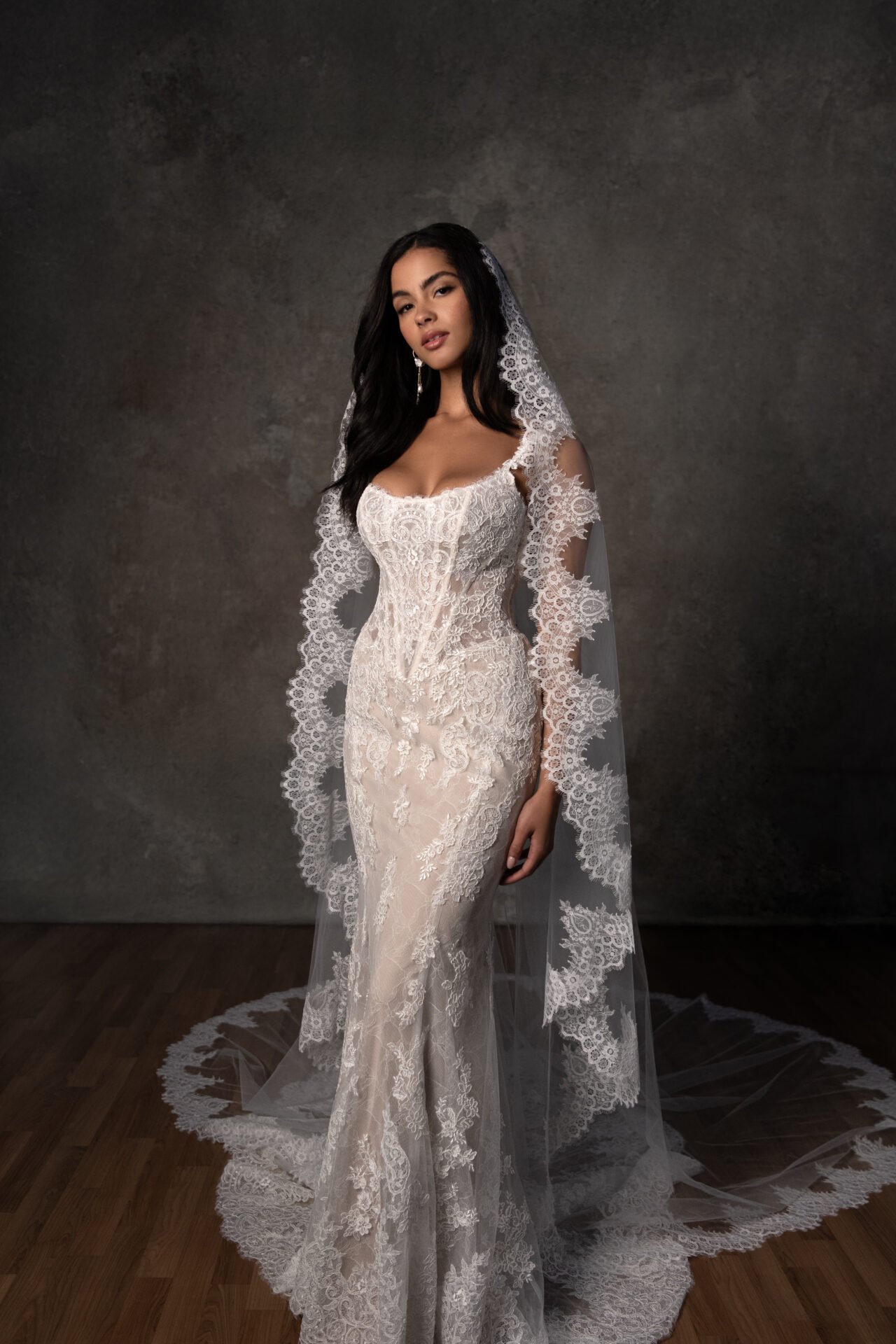 A woman in a wedding dress with lace details, wearing a long veil, standing on a wooden floor against a dark background.