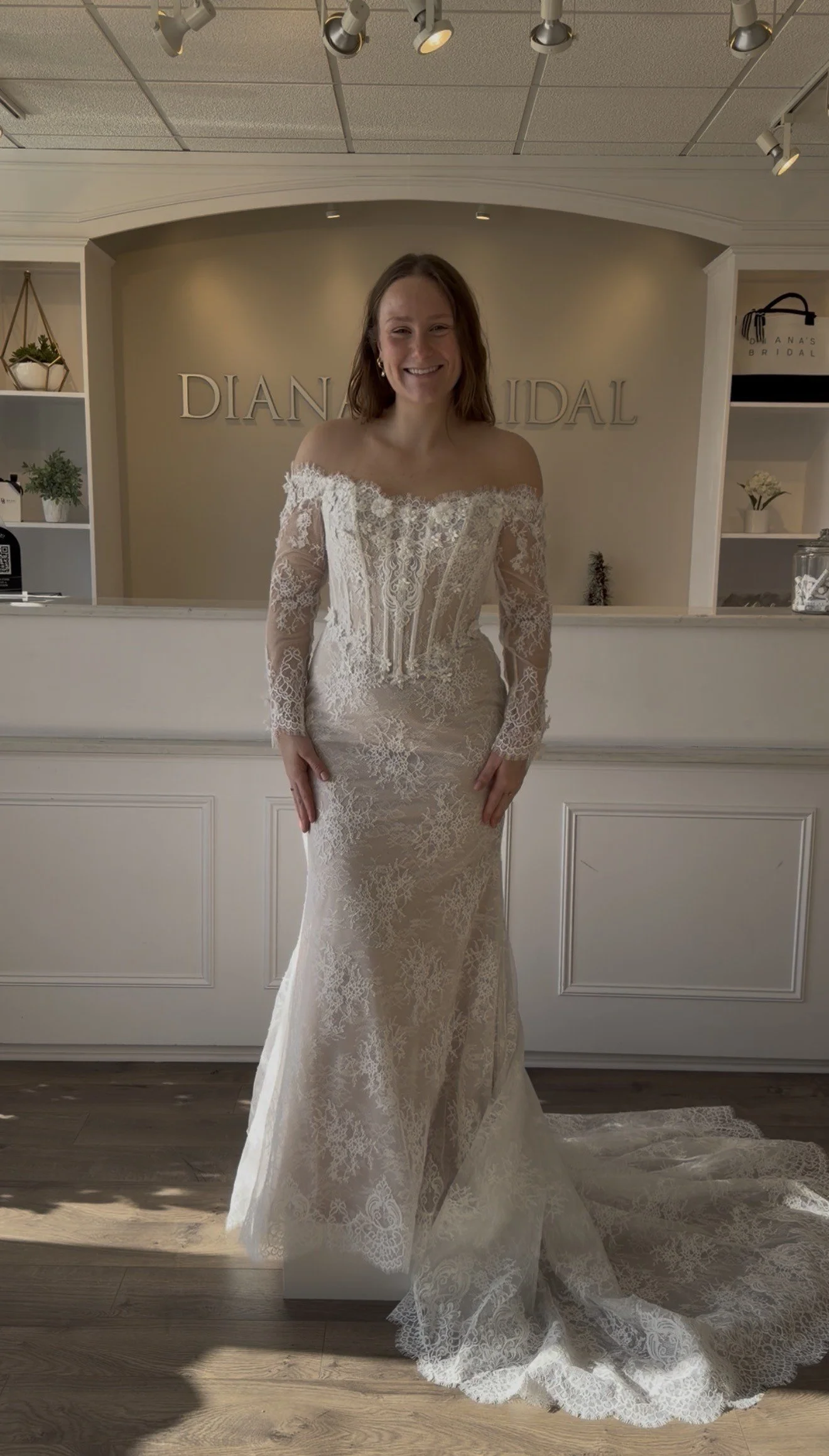 A woman in a white lace wedding dress standing inside a bridal boutique with a reception desk in the background. The woman is smiling and has shoulder-length brown hair.