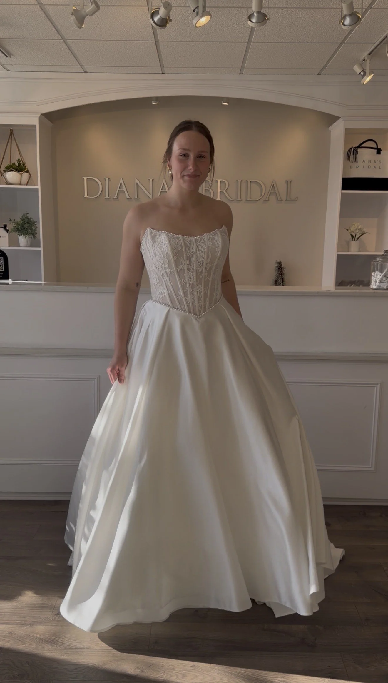 A woman trying on a wedding dress in a bridal boutique. The dress has a fitted, lace bodice and a full satin skirt. The boutique has a reception area with plants and decorations.