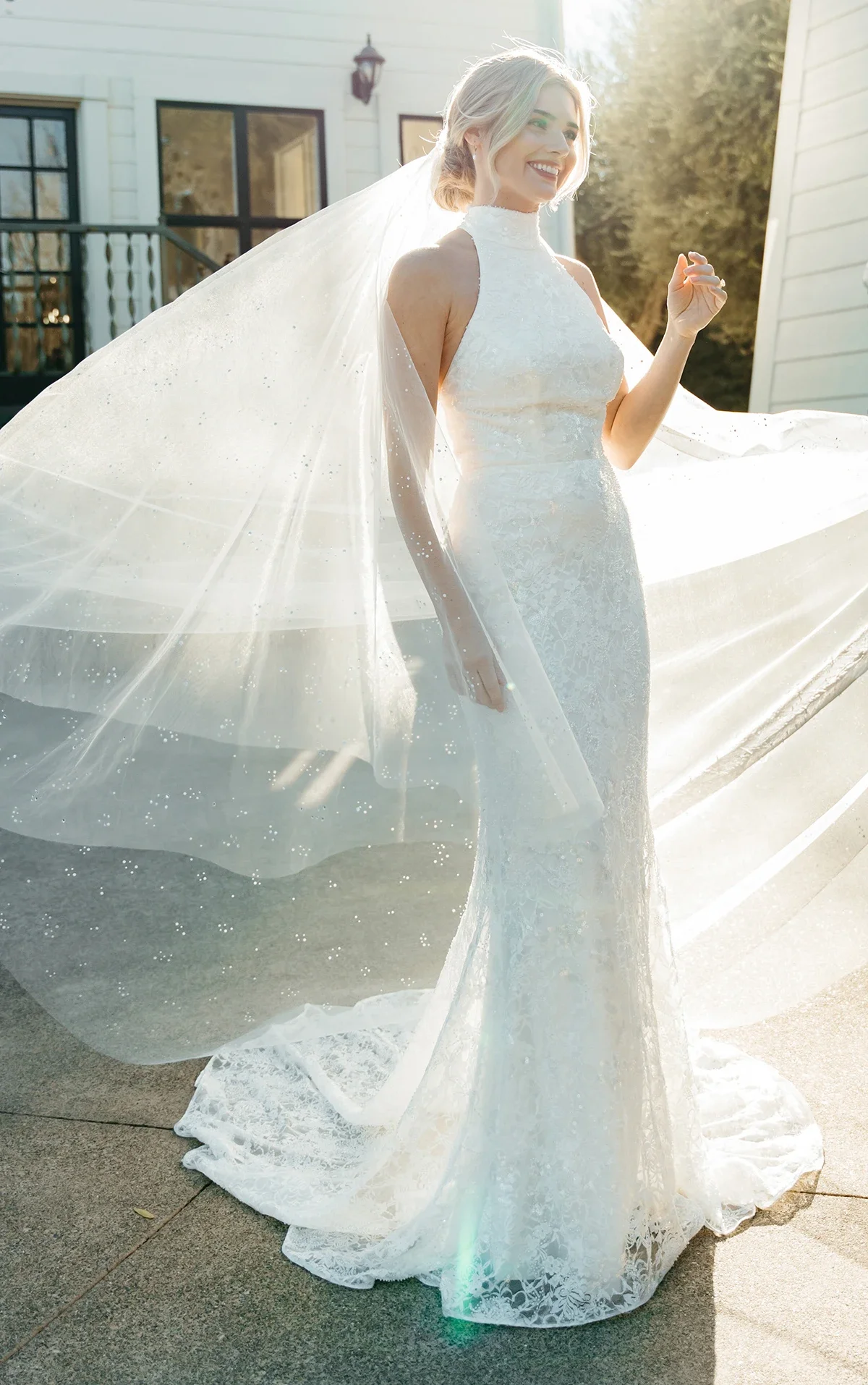 A smiling bride with light-colored hair in a lace wedding dress and flowing veil standing outdoors in front of a house during sunlight.