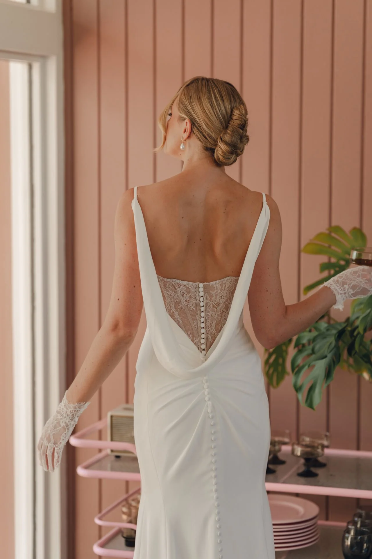 Back view of a woman in a white dress with lace details, standing indoors near a plant, with wooden panel walls.