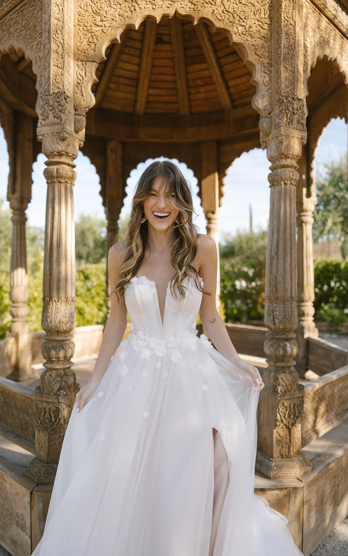 A woman in a white wedding dress with floral details smiling in front of an ornate wooden gazebo outdoors.