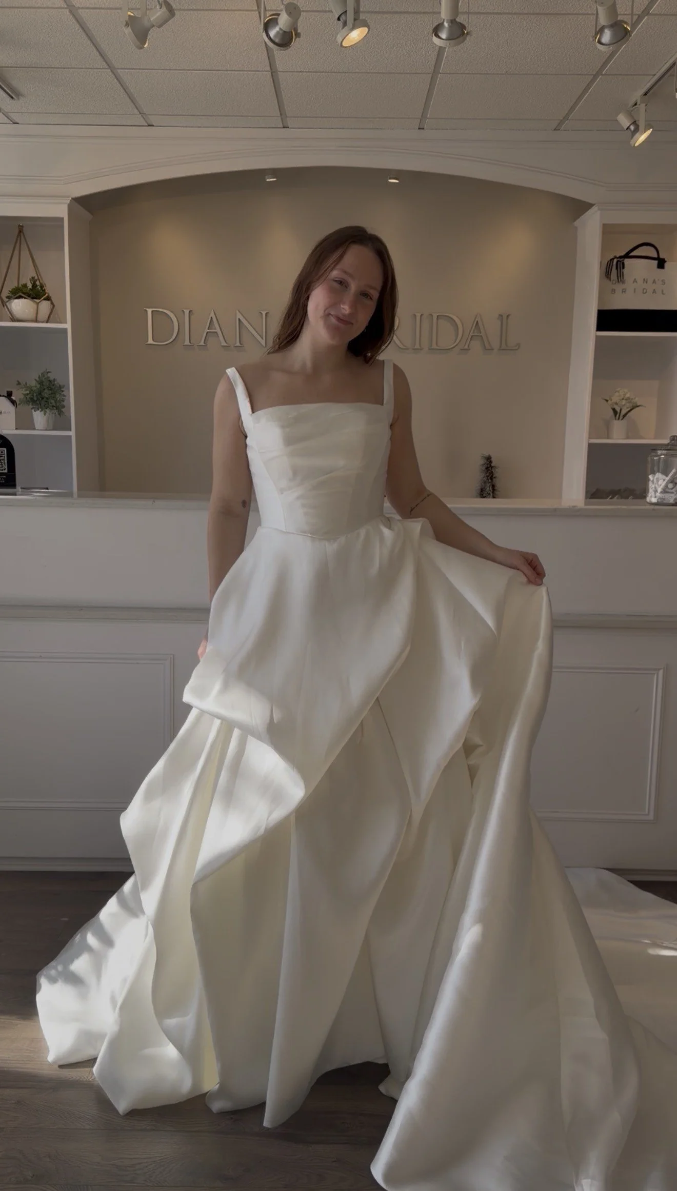 A woman trying on a white bridal dress inside a bridal shop, with the shop's sign 'DIANA BRIDAL' visible in the background.
