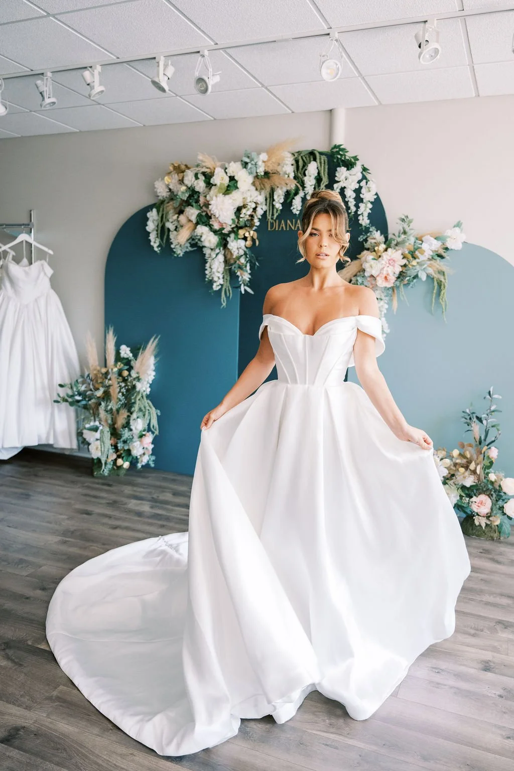 A woman trying on a white off-the-shoulder wedding dress in a bridal boutique, with floral decorations and a display rack with more dresses behind her.