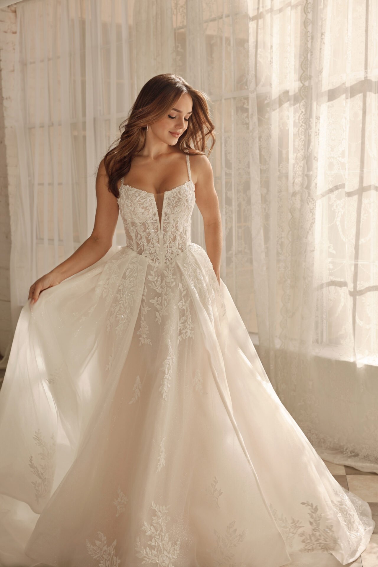 A woman in a wedding dress with lace details, standing in front of a window with sheer curtains, holding the skirt of her dress
