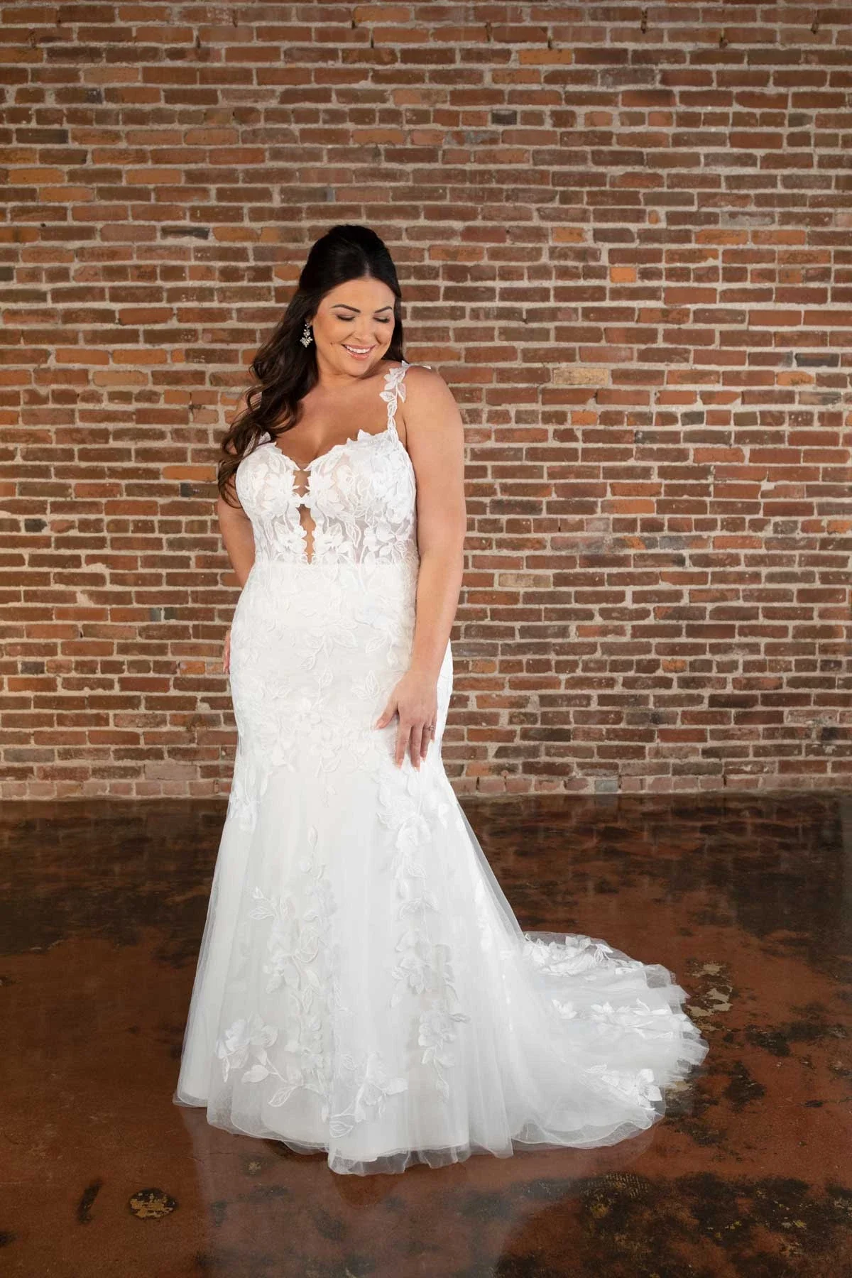 A bride in a white lace wedding dress with floral embroidery, standing against a brick wall, smiling and looking down.