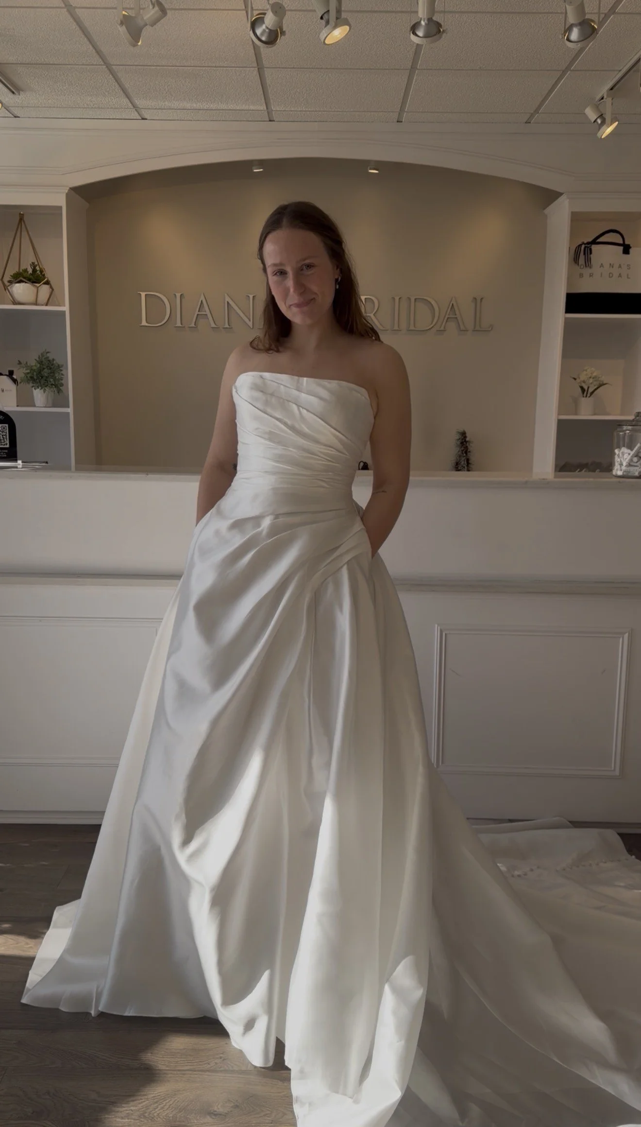 A woman trying on a white wedding dress in bridal boutique with the sign 'Diana Bridal' in the background.