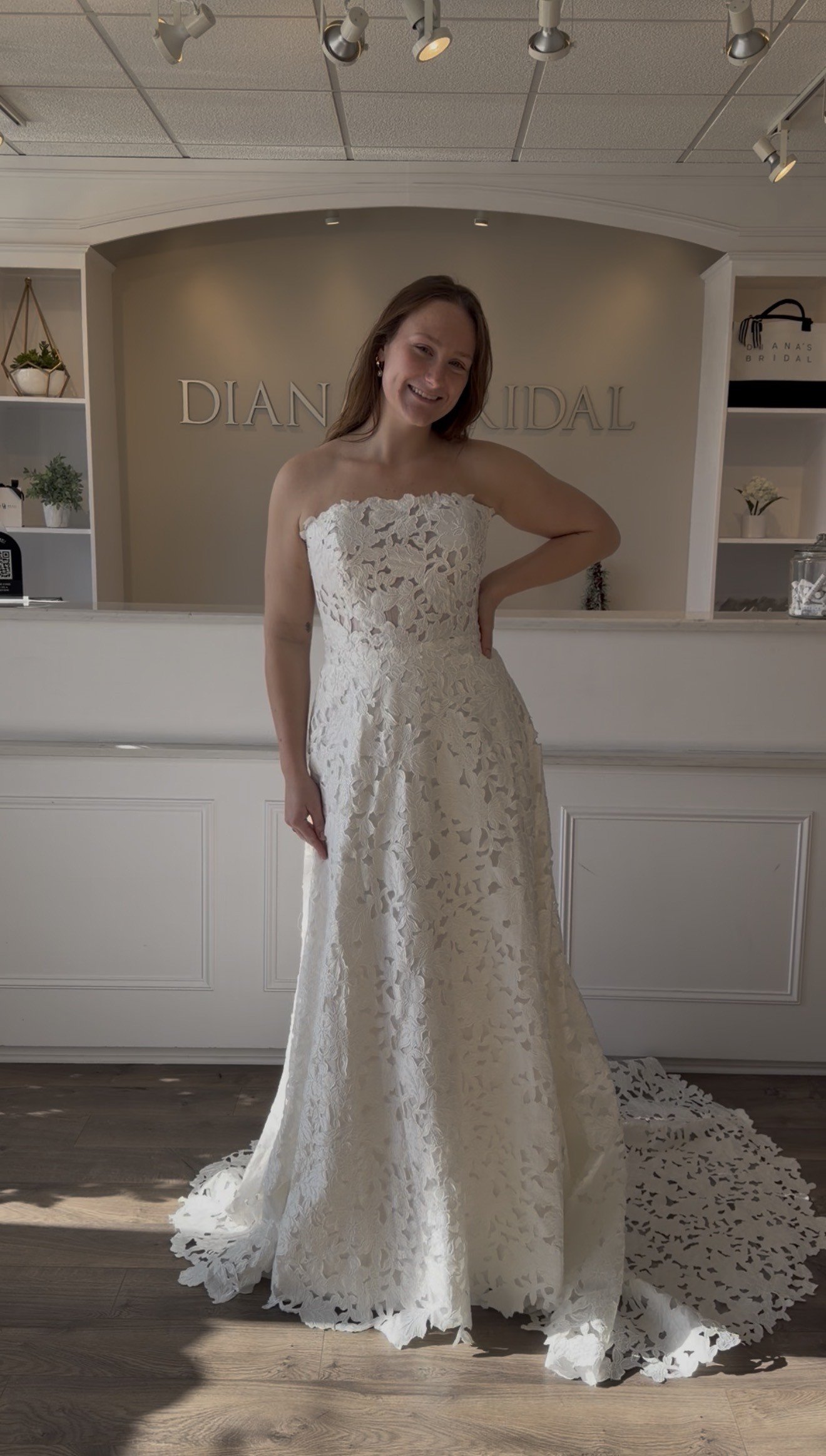 A woman trying on a white lace wedding dress inside a bridal boutique, smiling at the camera.