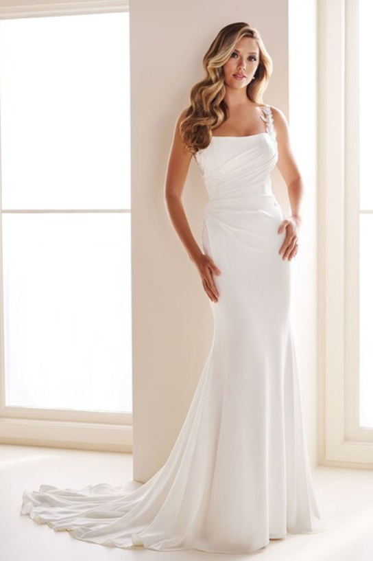 A woman in a white wedding dress standing near a window with sunlight coming through.