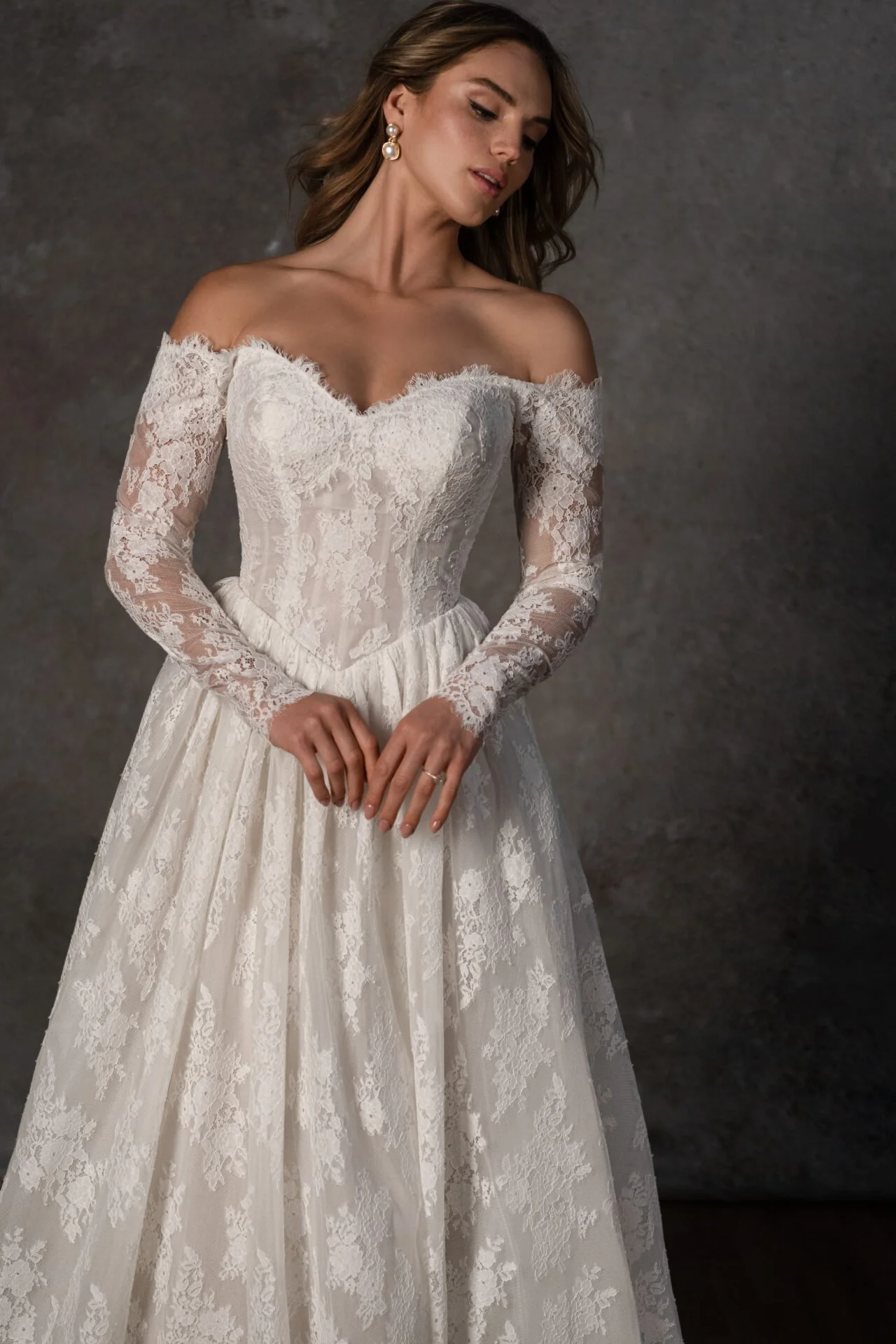 A woman in a white lace off-shoulder wedding dress, with long lace sleeves, standing against a gray backdrop, looking downward.