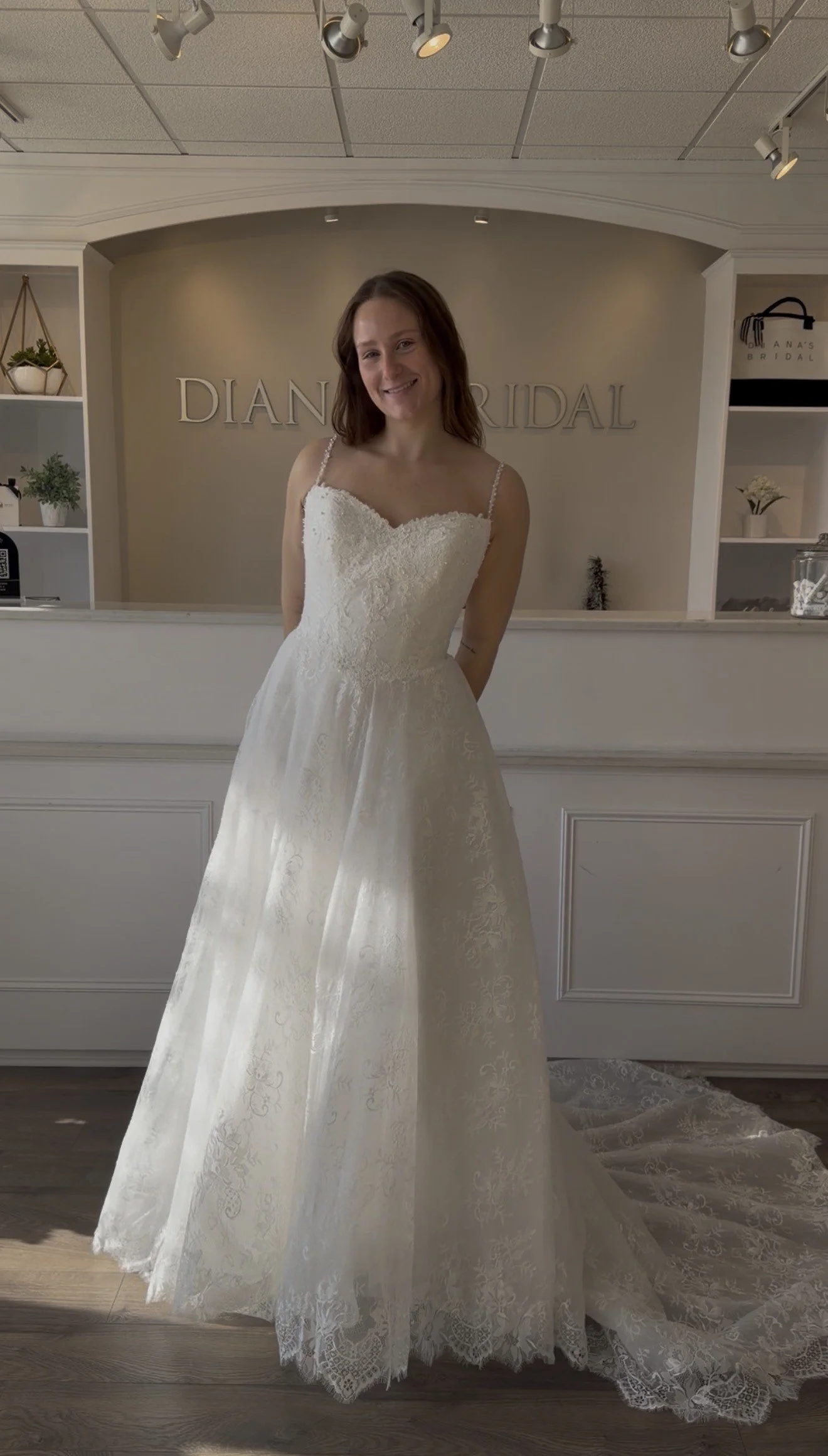 A woman in a white lace wedding dress stands inside a bridal boutique with a sign that reads 'DIANA'S BRIDAL' behind her. She is smiling and has her hands behind her back.