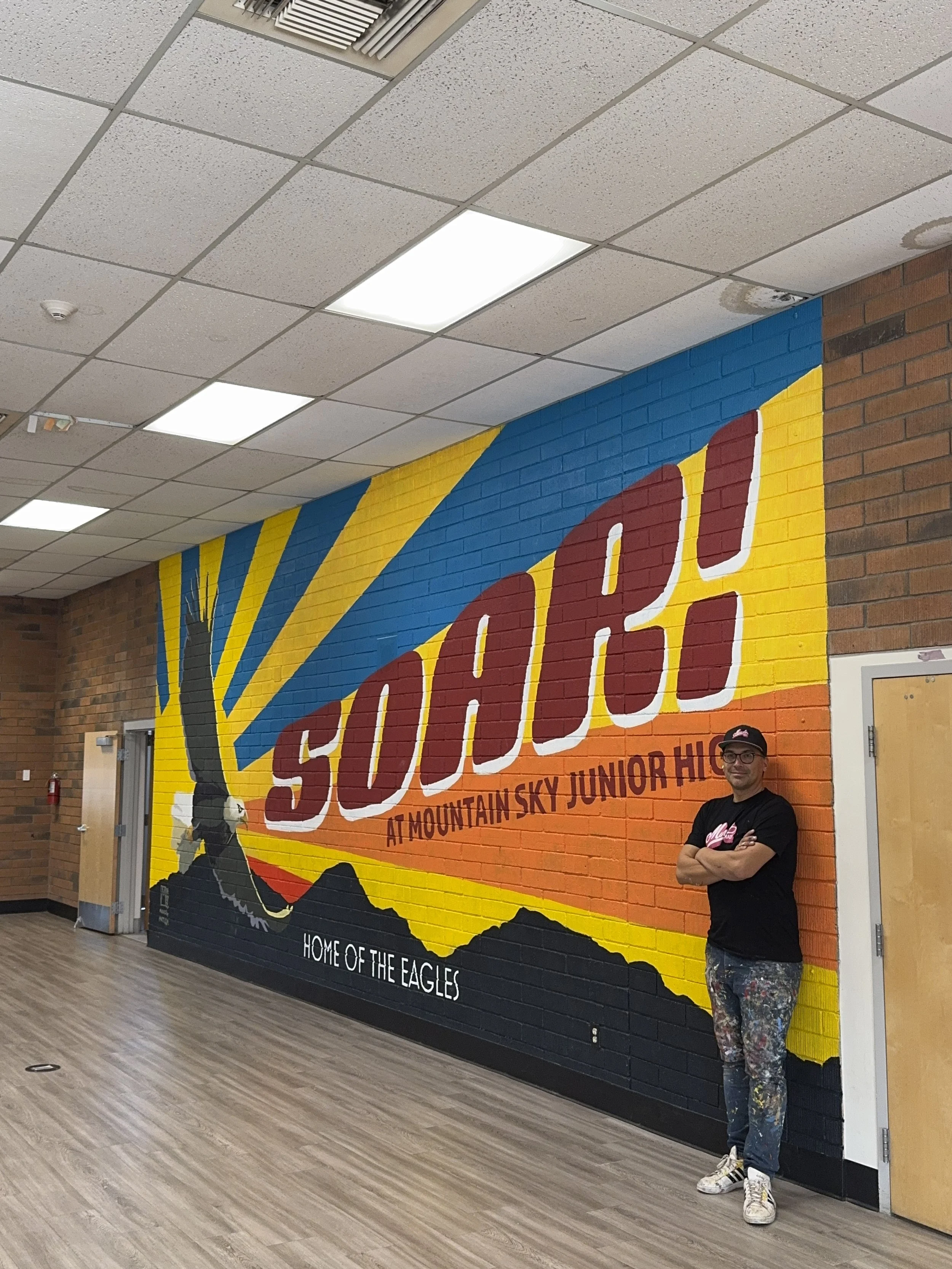 mural artist standing in front of a large mural on a brick wall at a school