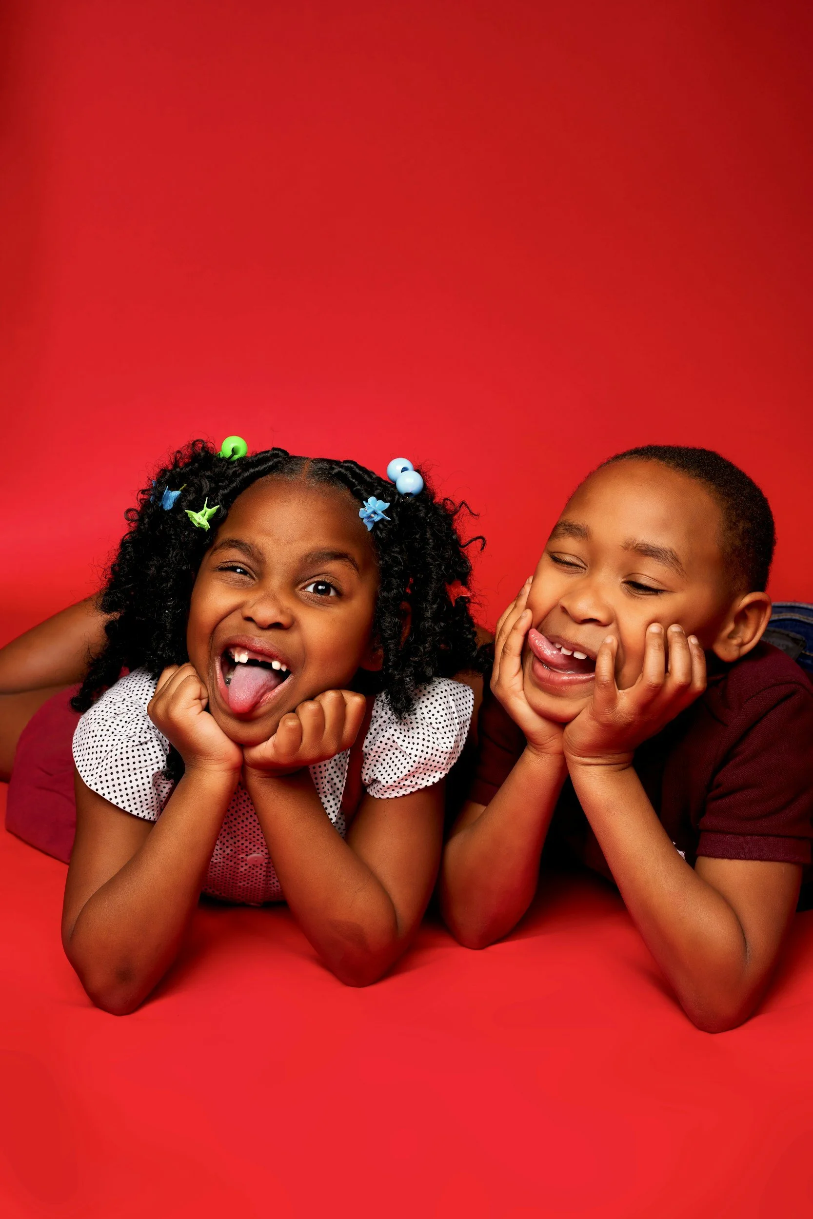 Two children lying on a red background, smiling and making silly faces. The girl on the left has curly hair with colorful beads, and the boy on the right has short hair.