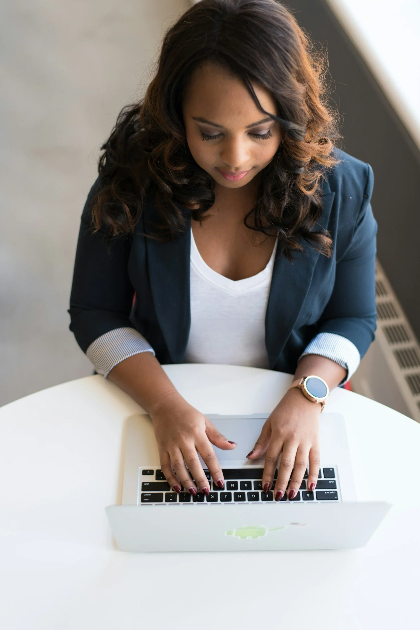 A young woman with dark wavy hair sitting at a white table, using a silver laptop with a green logo, wearing a navy blazer over a white top, with a smartwatch on her left wrist and manicured nails.