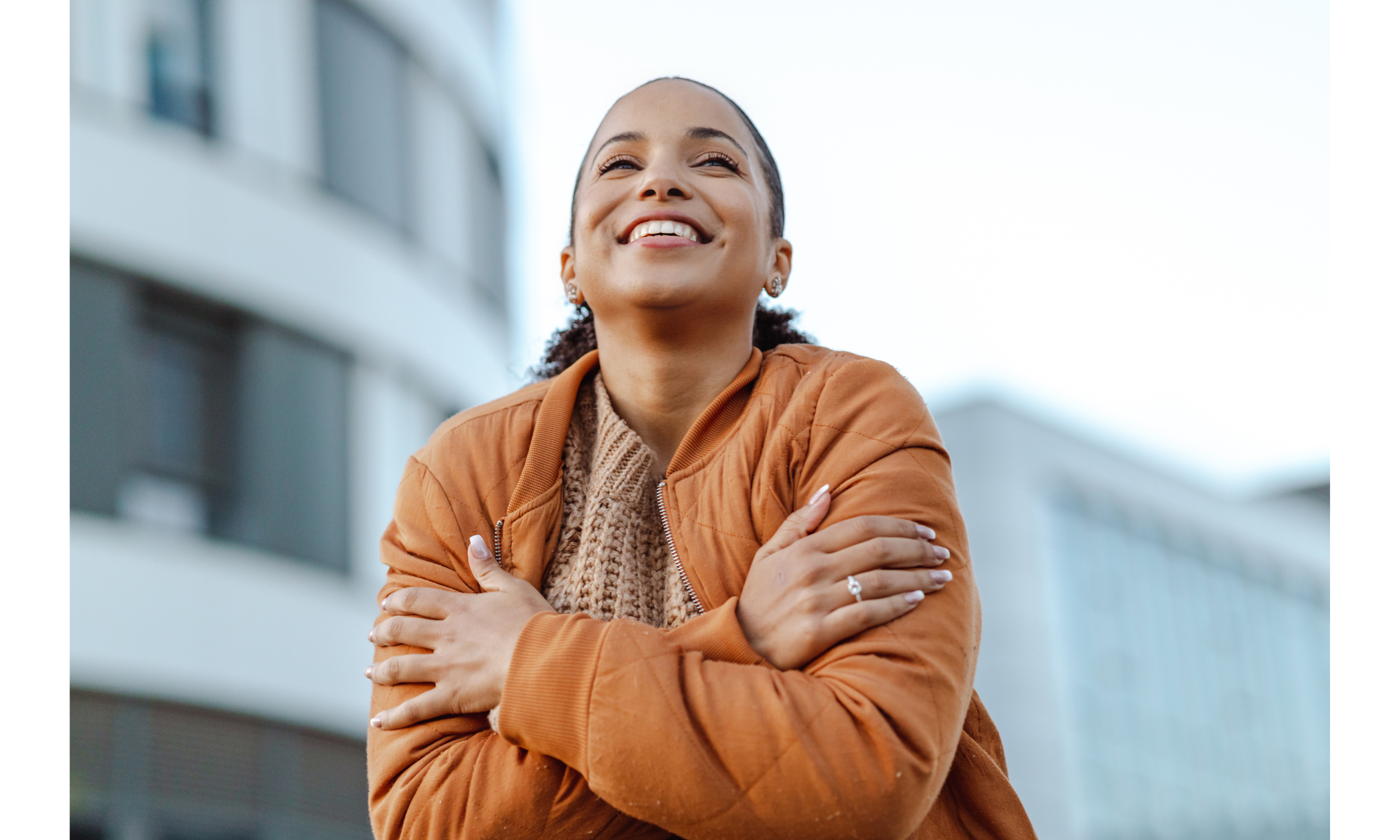 A woman smiling outdoors, wearing a brown jacket and crossing her arms.