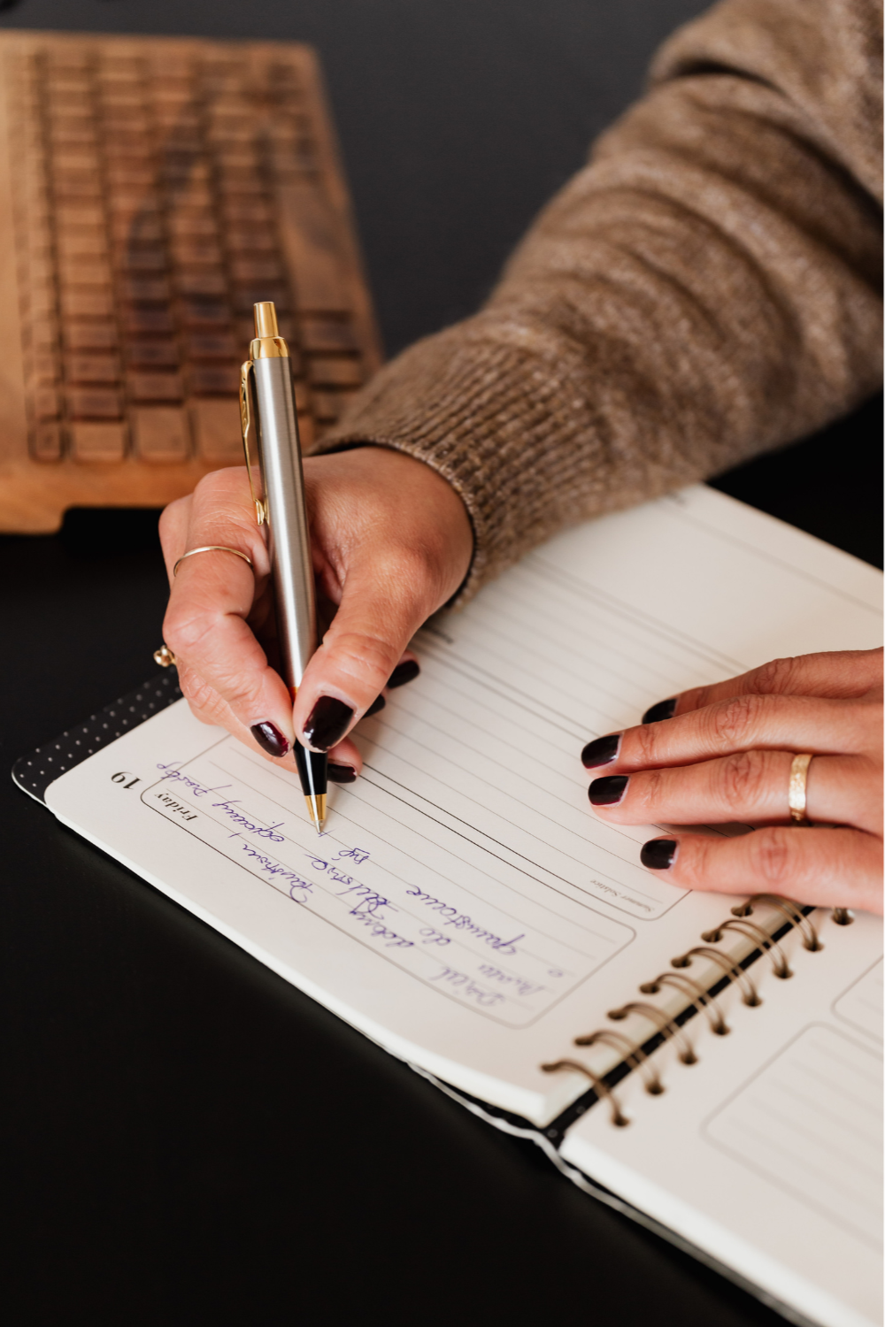 A person with dark nail polish writing in a planner with a black ballpoint pen, wearing a beige knit sweater, on a black surface. Part of a wooden keyboard is visible in the background.