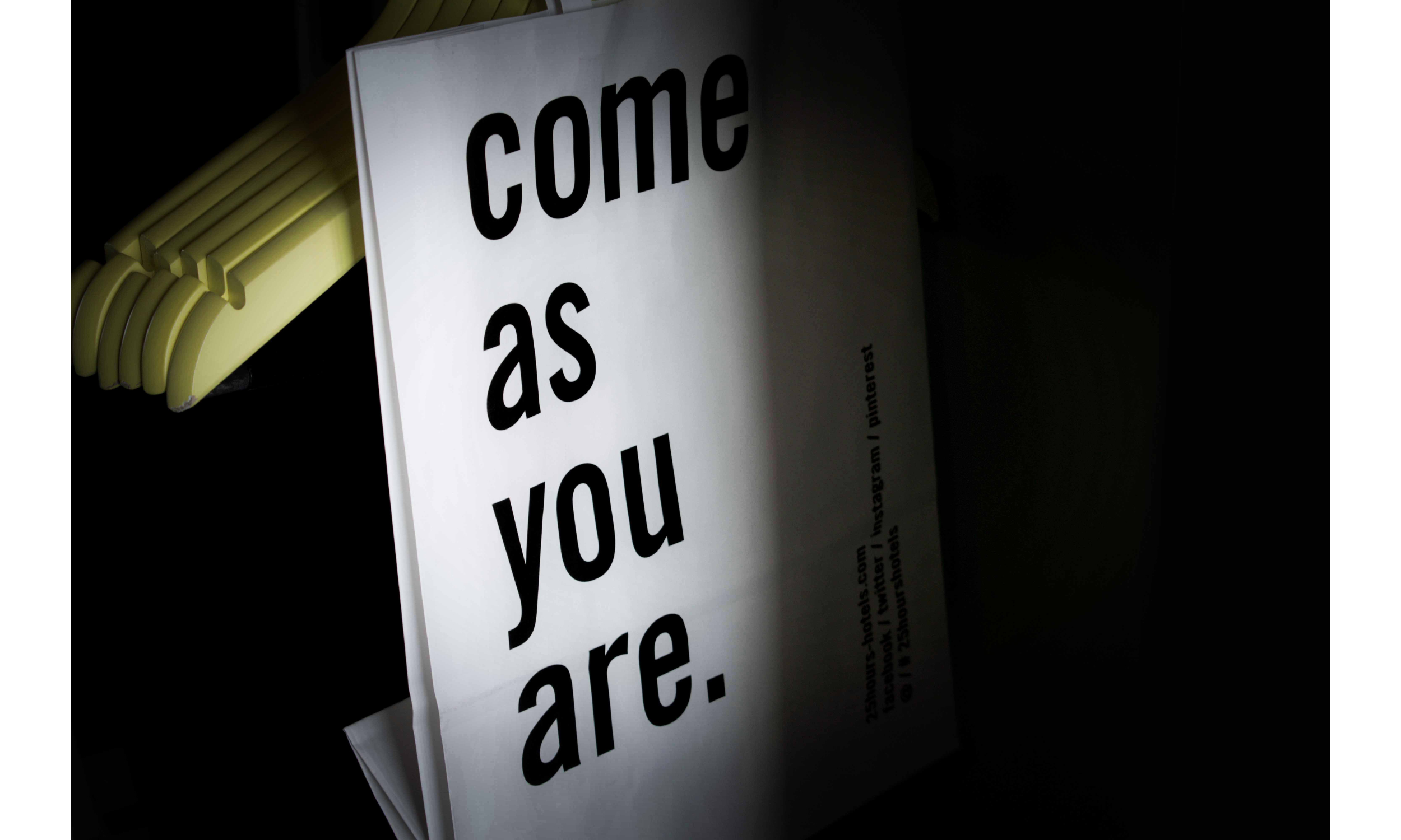 Close-up of a white sign with bold black text reading 'come as you are' on a dark background, partially covered by yellow coat hangers.