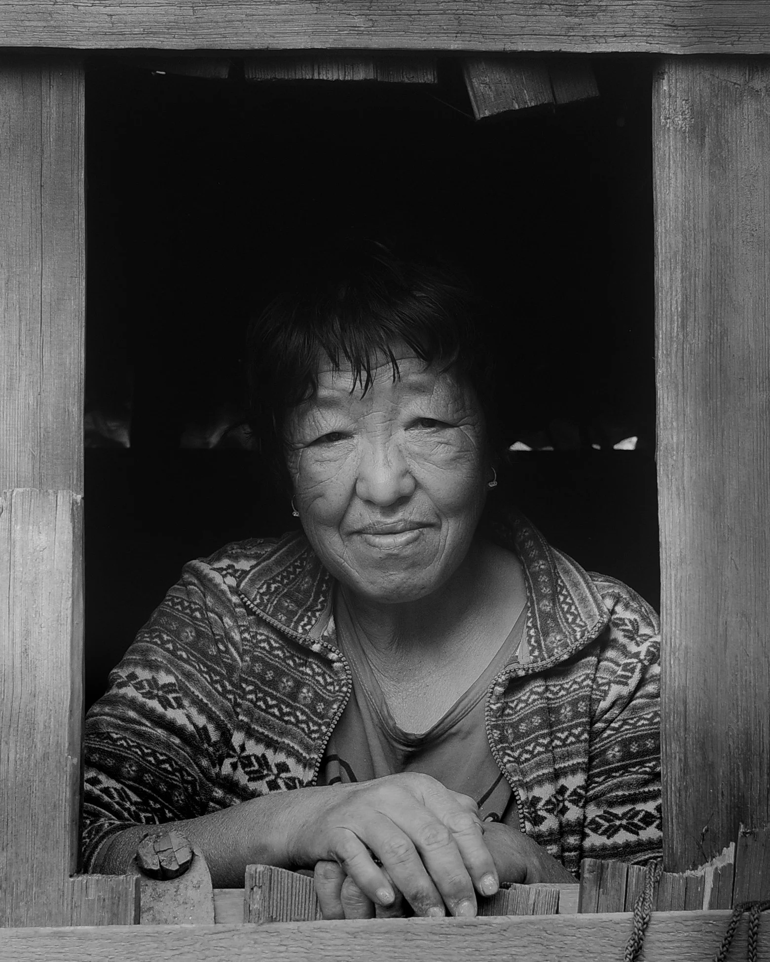 An elderly woman with short dark hair and a patterned sweater, smiling, looking out from a square opening in a wooden structure.