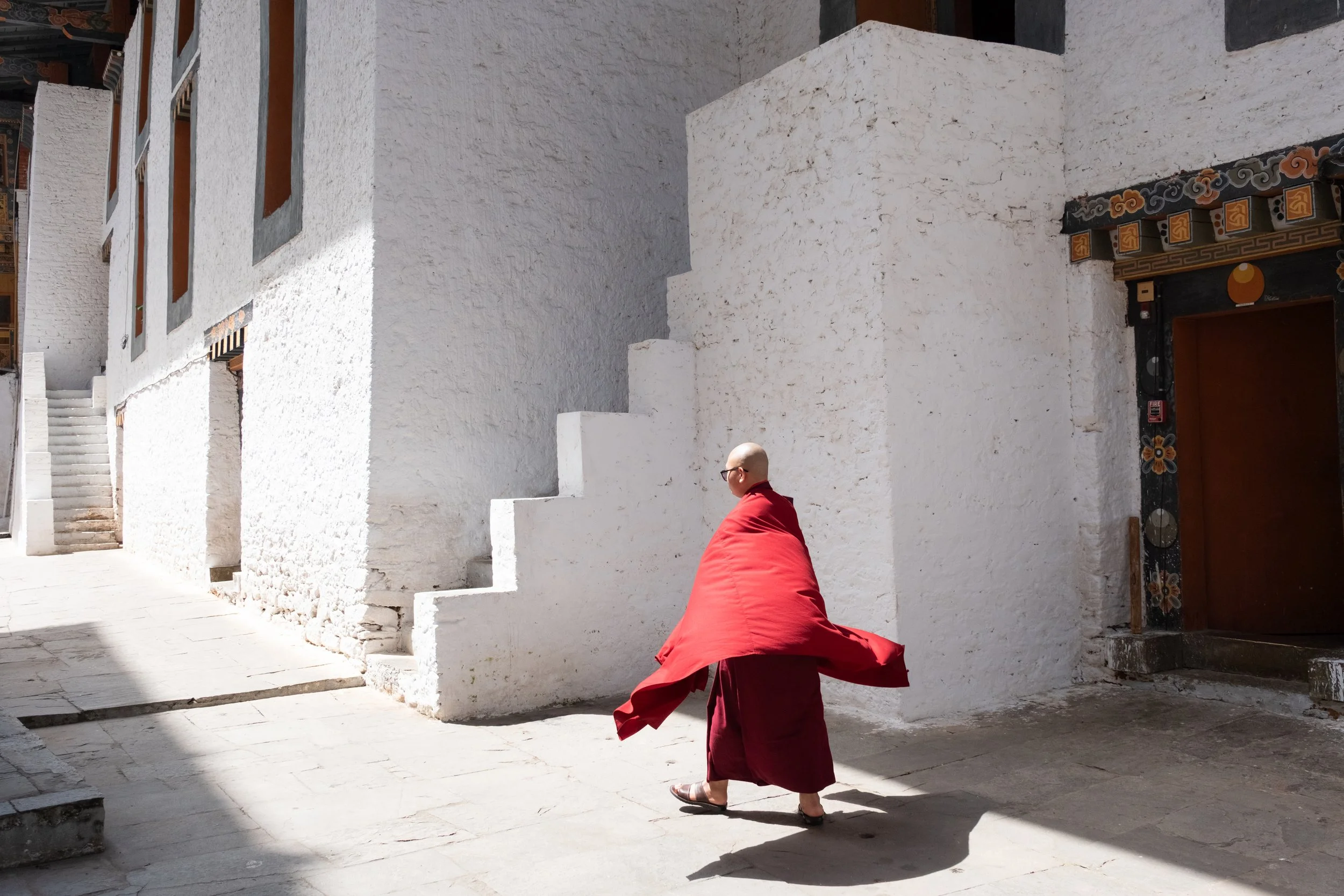 A person wearing a red jacket and sandals walking along a white stone building with traditional decorative trim near the door in a sunny outdoor setting.