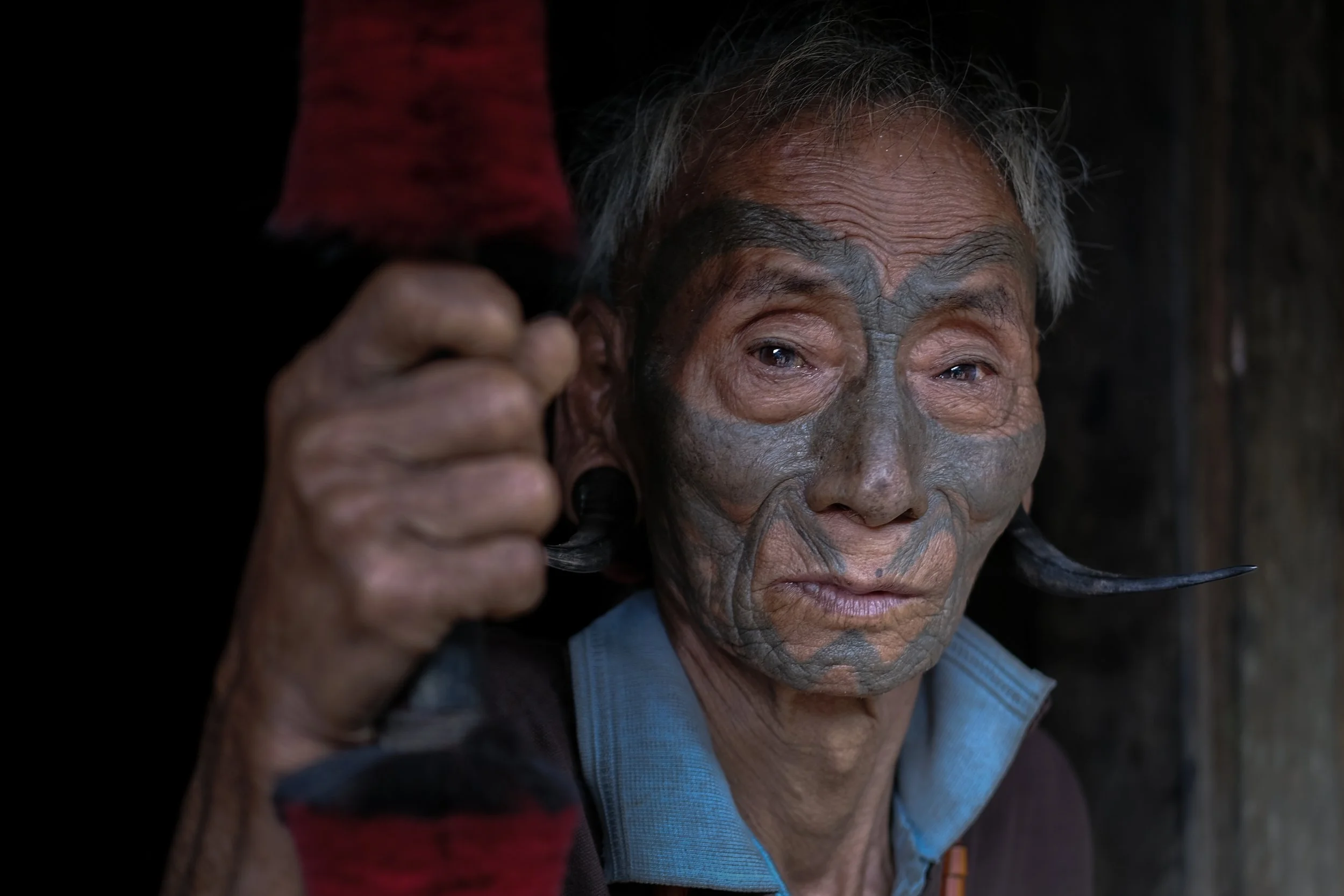 Close-up portrait of a Konyak elder with facial tattoos, holding a red-wrapped spear handle beside his face and looking directly at the camera.