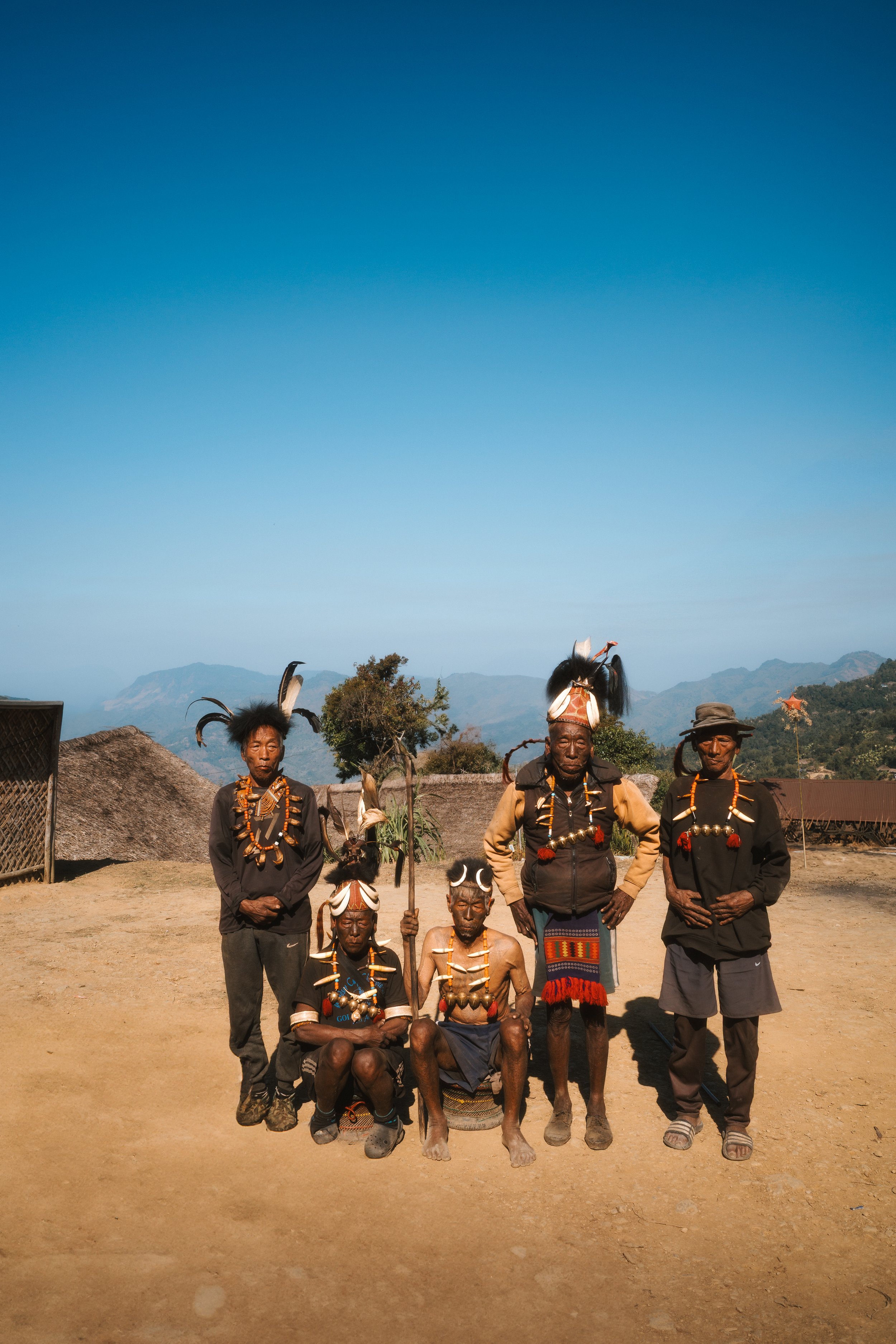 Five Konyak men in traditional dress, feathered and horned headdresses, and beaded necklaces standing and sitting on a hillside overlooking the mountains.