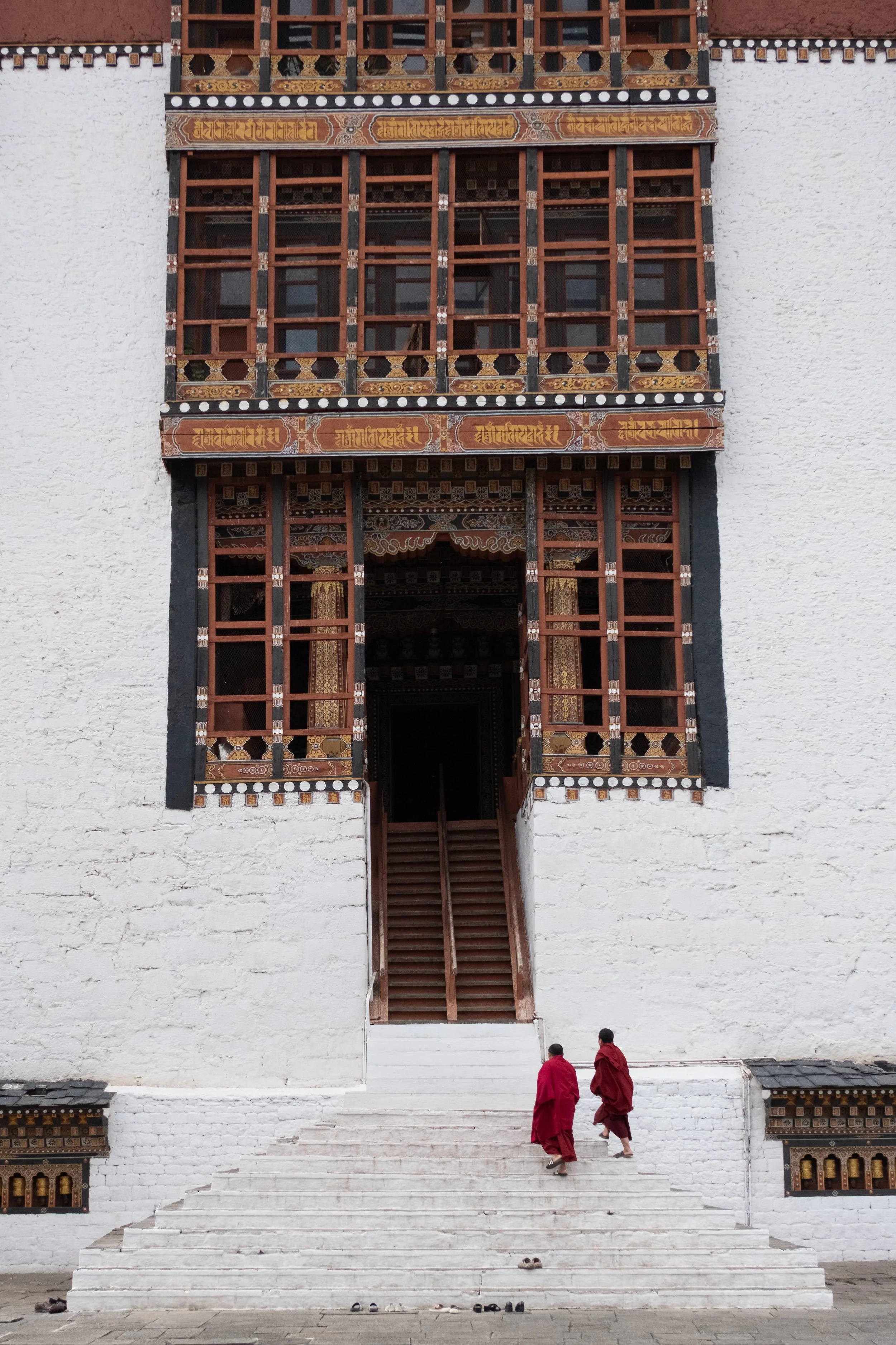 Two monks in red robes walking up white stone steps toward the entrance of a traditional Tibetan Buddhist monastery. The building is white with intricate wooden window frames and colorful decorative elements.