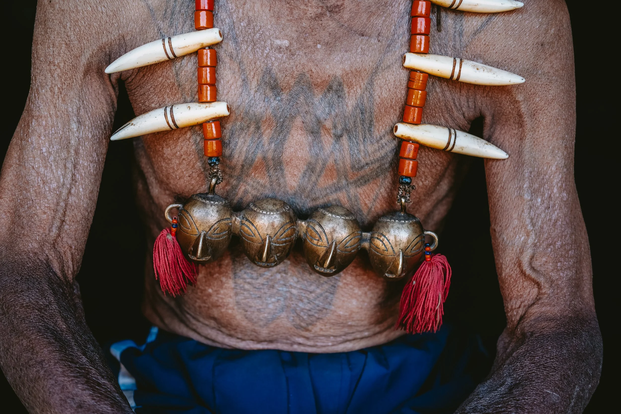 Close-up of a tattooed Konyak man's chest, wearing a necklace of brass bells, bone ornaments and red tassels.