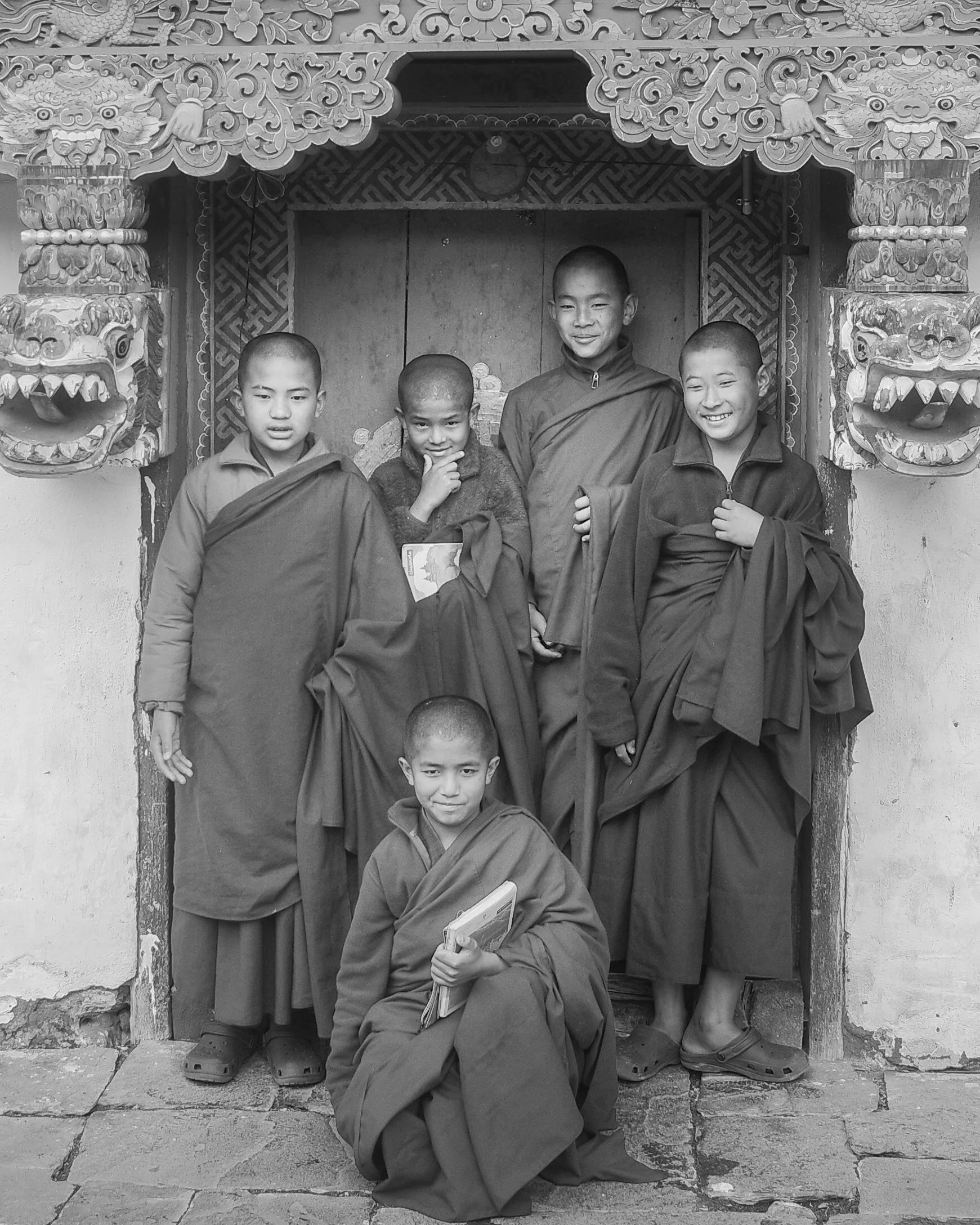 A black and white photo of five young monks, four standing behind a door frame and one kneeling in front, in front of an ornate wooden door in a traditional temple or monastery. The two dragons carved on either side of the door frame are visible, wit