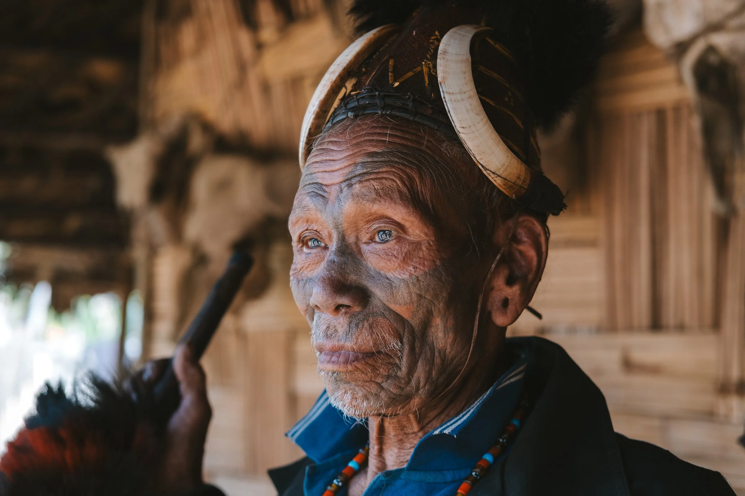 Close-up of an elderly person with detailed wrinkles, wearing traditional jewelry and headgear, inside a rustic wooden structure.