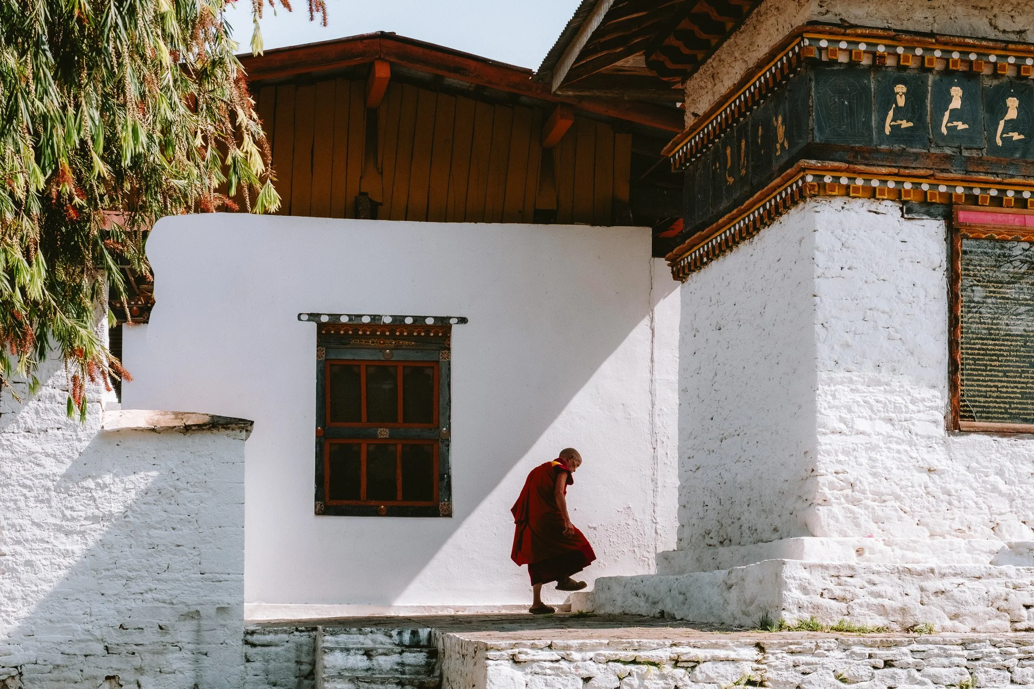 A person in red robes walking down white stone stairs outside a building with white walls, a window, and decorative trim under the roof in a sunny setting.