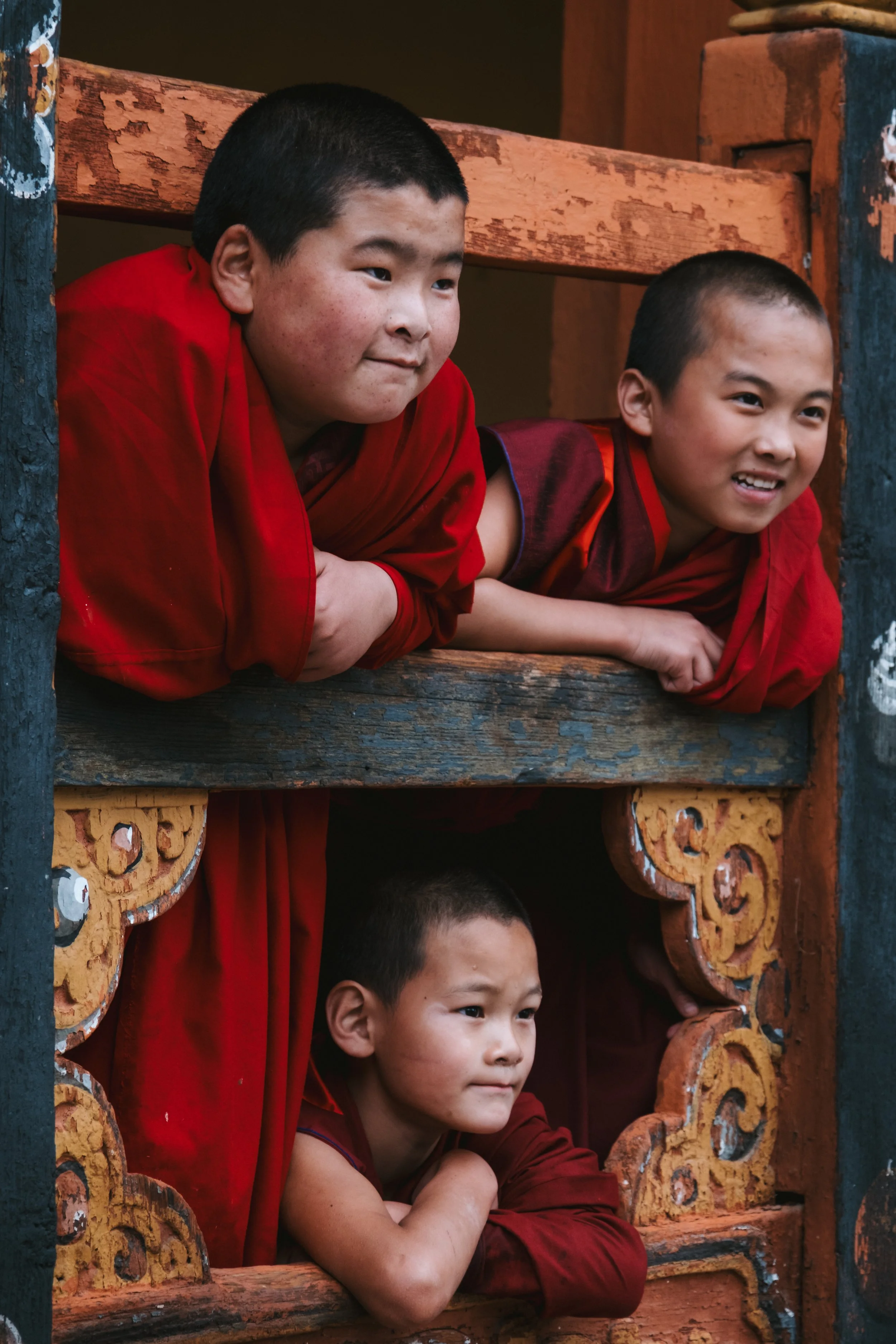 Three young boys dressed in traditional red robes are leaning out from the wooden balcony of a building with ornate orange and black carvings, looking down at something below. They appear to be in a Buddhist monastery or similar cultural setting.