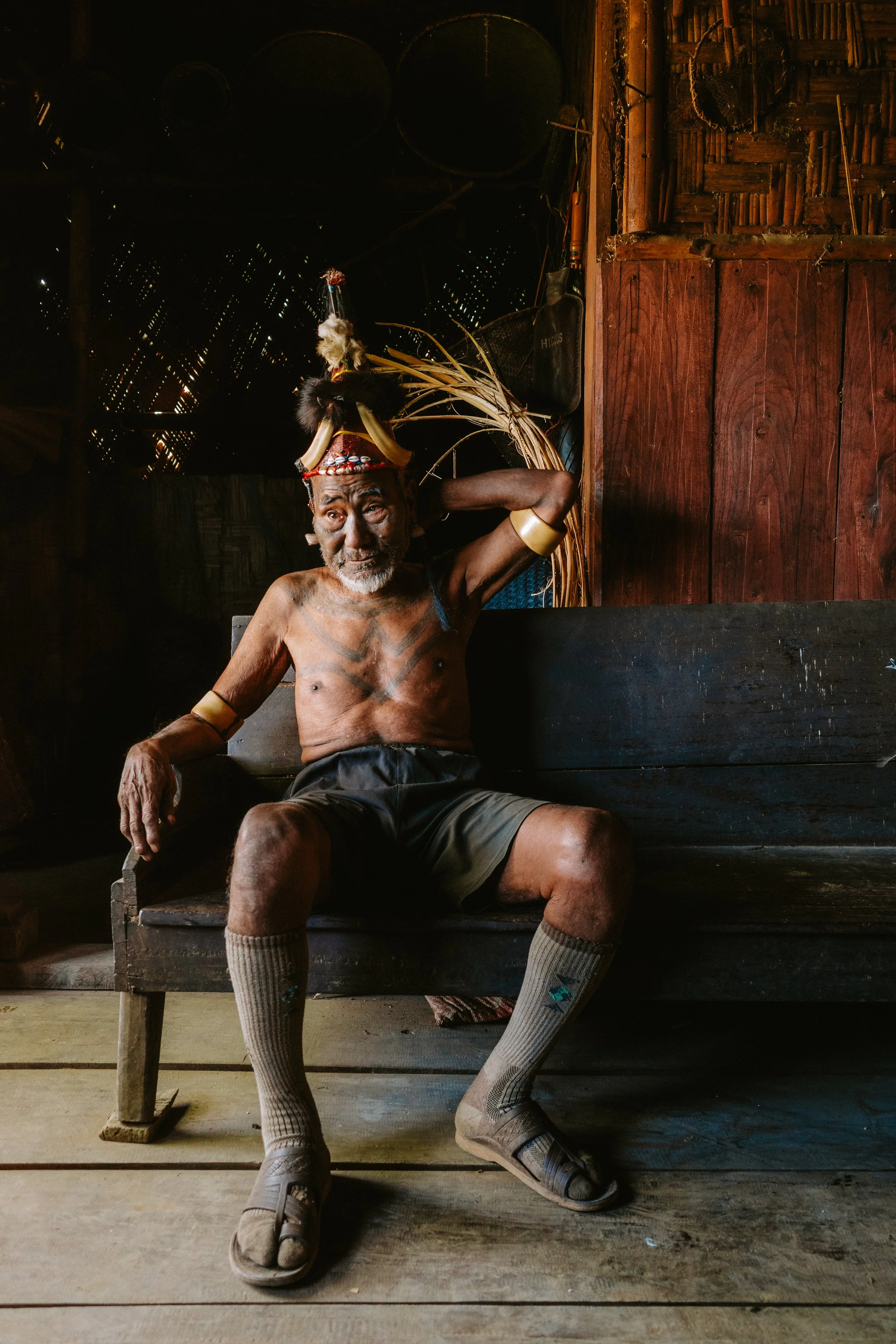 A shirtless Konyak elder wearing a feathered headdress and arm bands, seated on a wooden bench inside a wooden house.
