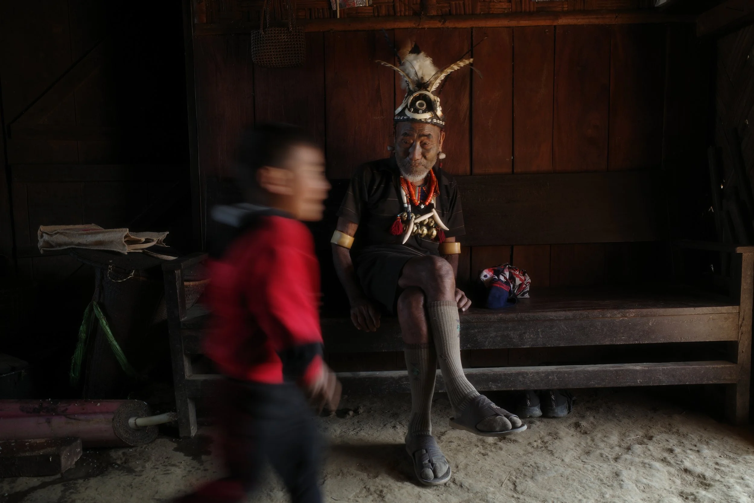 An elderly man wearing traditional attire with a feathered headdress sits on a wooden bench inside a dimly lit wooden hut. A blurred child in red walks past in the foreground.