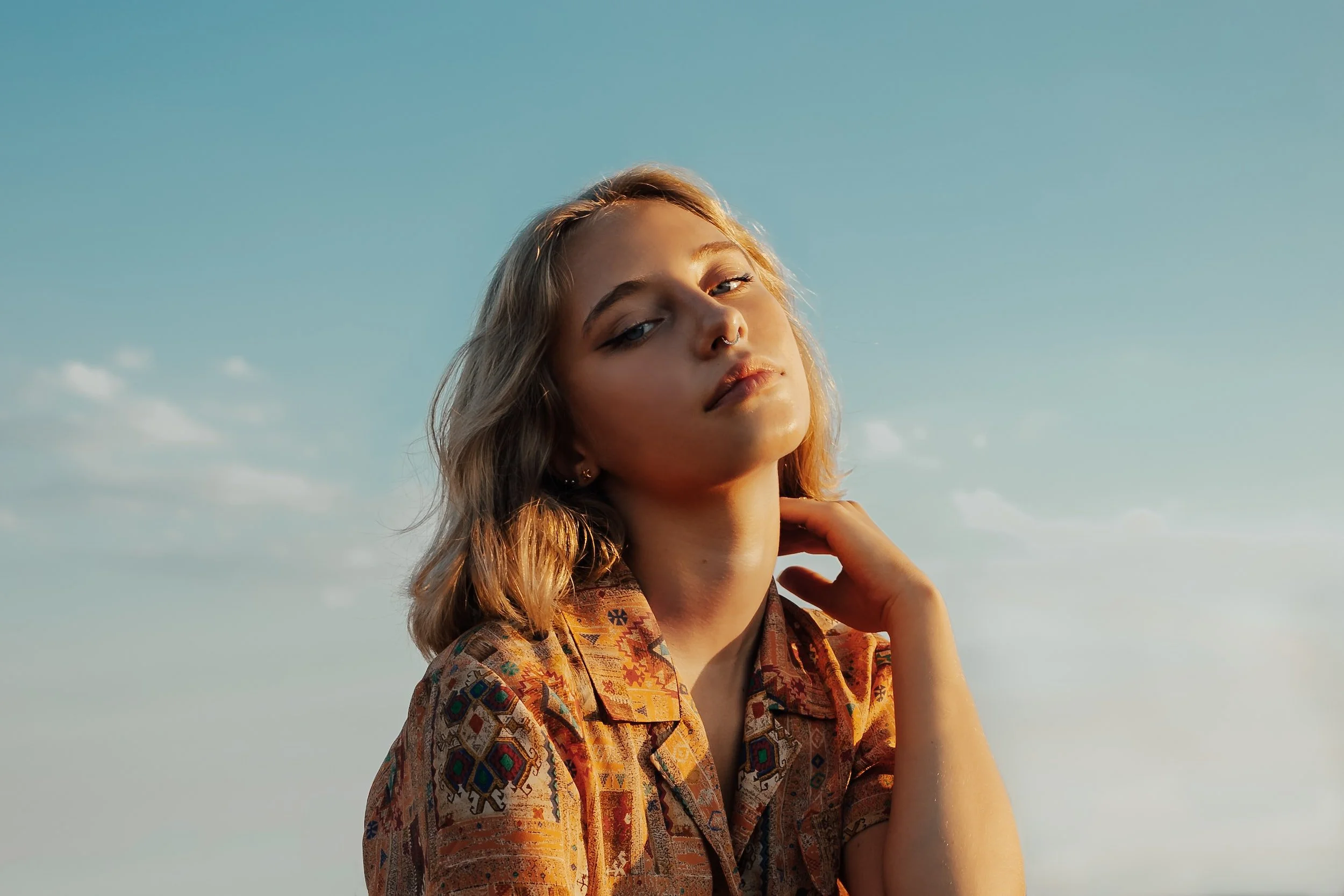 A young woman with blonde hair and a septum piercing standing outdoors in the sunlight, wearing a colorful patterned shirt, and looking at the camera with a serene expression, against a backdrop of a blue sky with a few clouds.