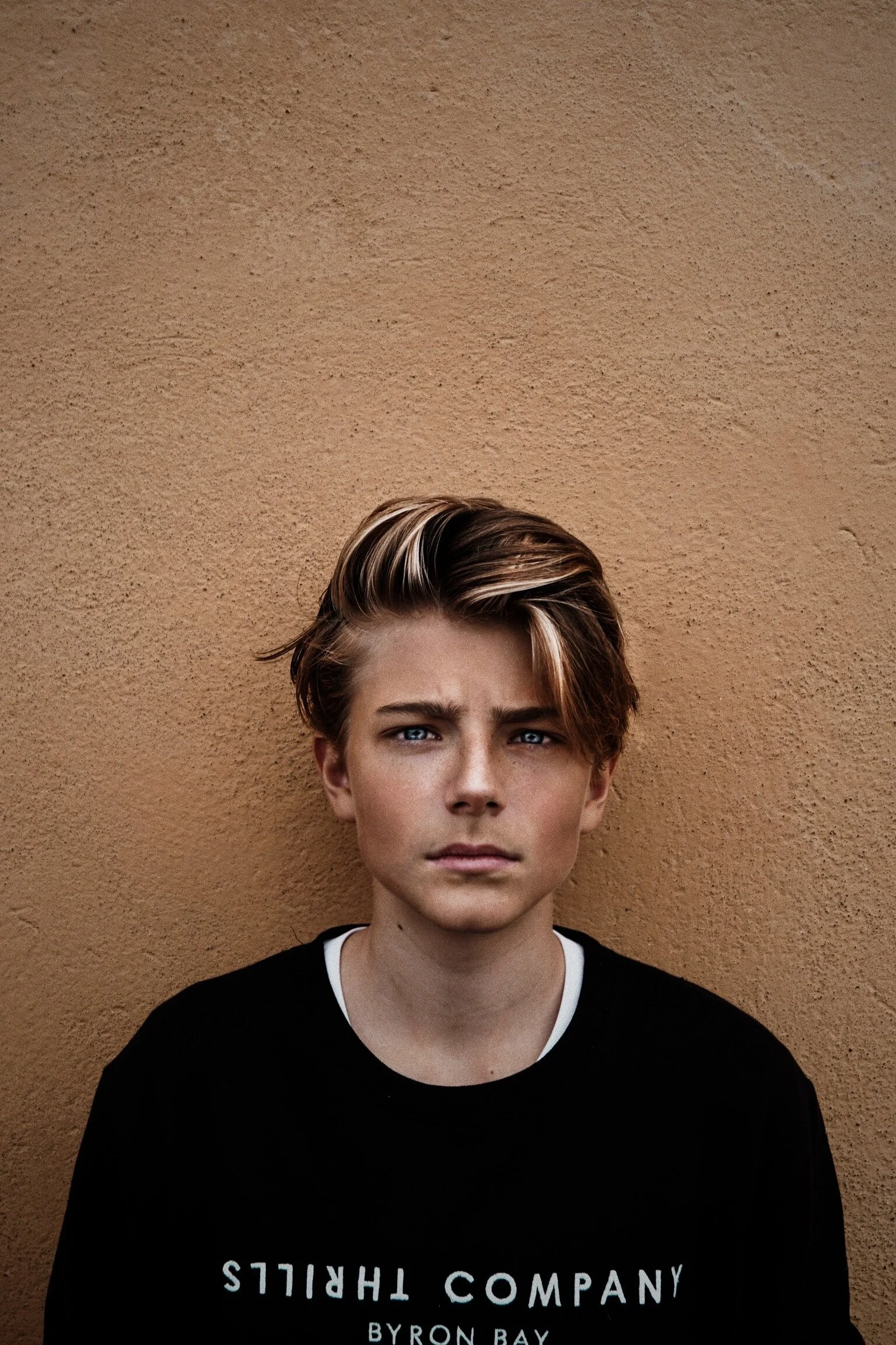 Portrait of a young man with styled blonde hair, blue eyes, and wearing a black T-shirt with white text, standing against a textured tan wall.