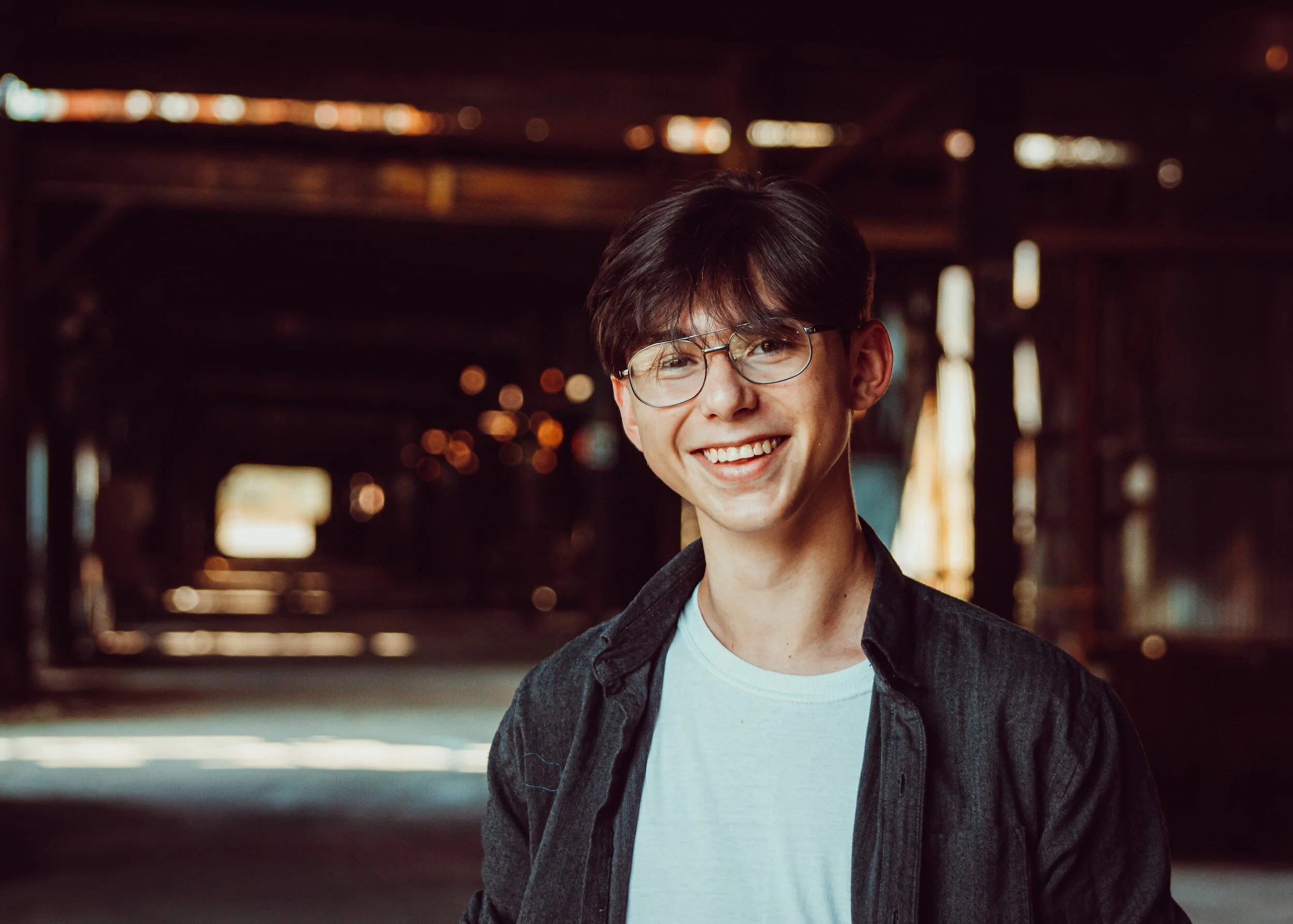 A young man with dark hair, glasses, and a big smile, wearing a black jacket and white t-shirt, standing inside a dimly lit industrial building or warehouse with blurred background lights.