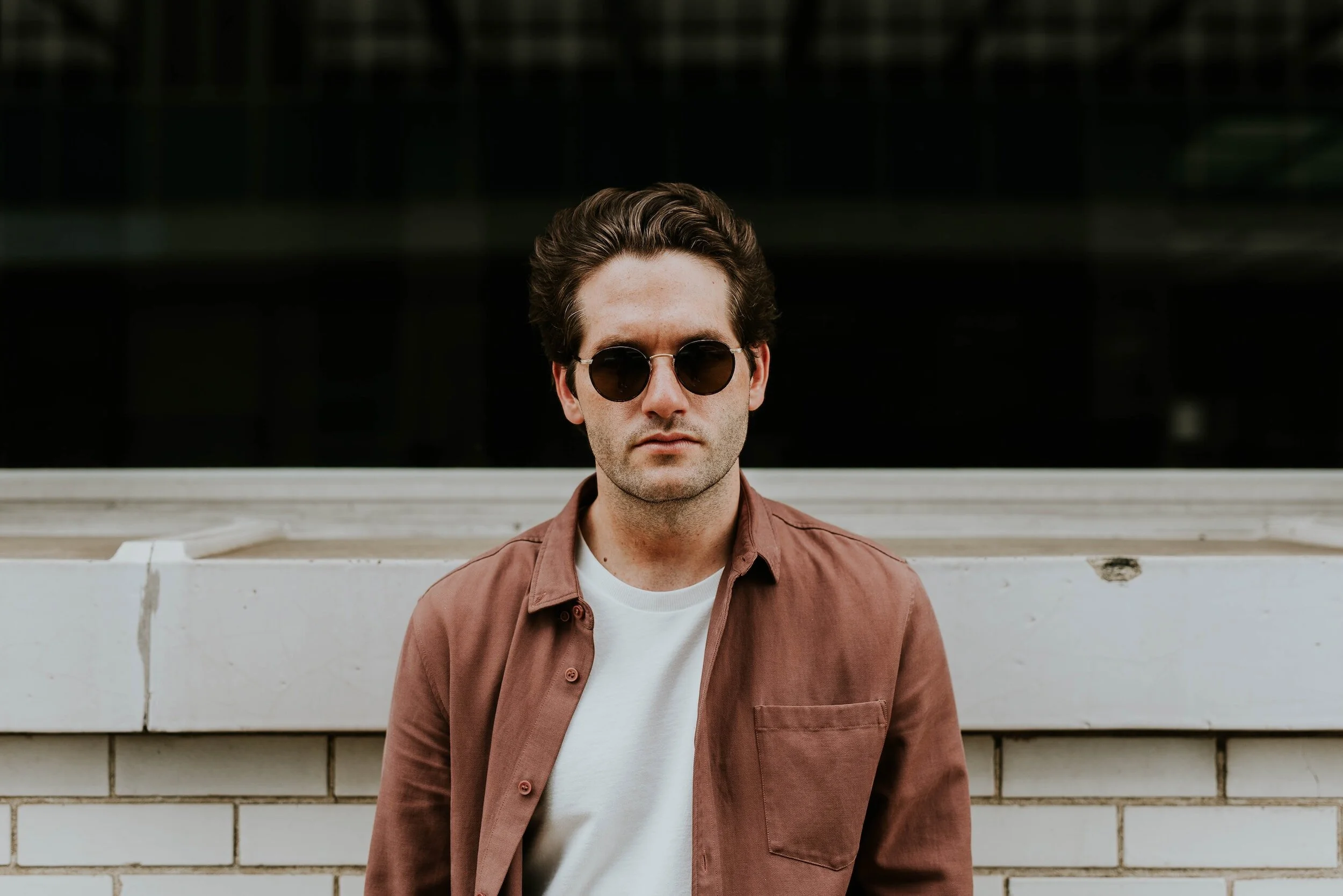 A young man with brown hair wearing round sunglasses, a brown button-up shirt over a white t-shirt, standing outdoors in front of a brick wall with a dark building behind him.