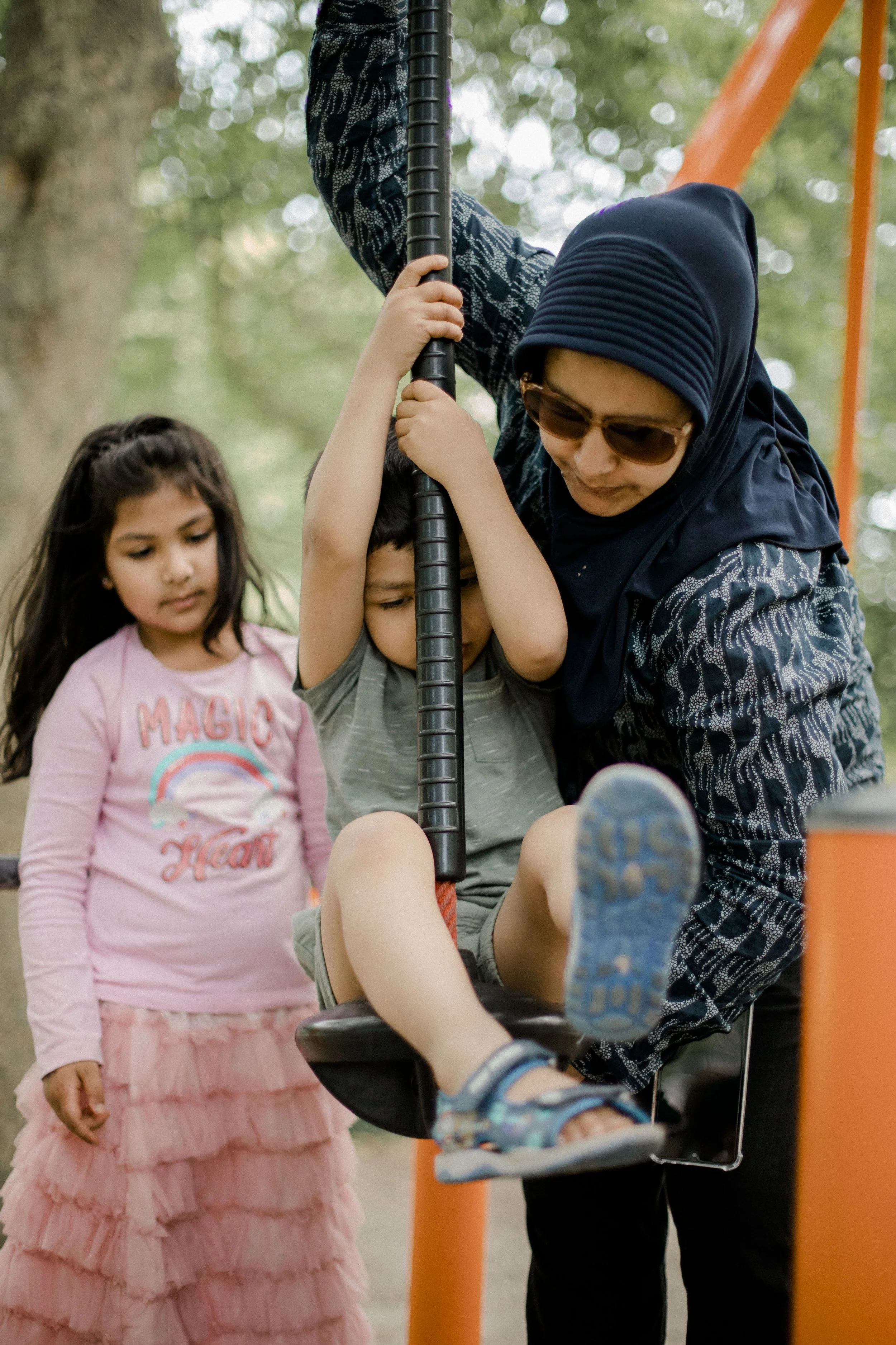 A woman wearing sunglasses and a navy blue hijab helps a young boy on a swing at a park, with a girl in a pink dress watching nearby.