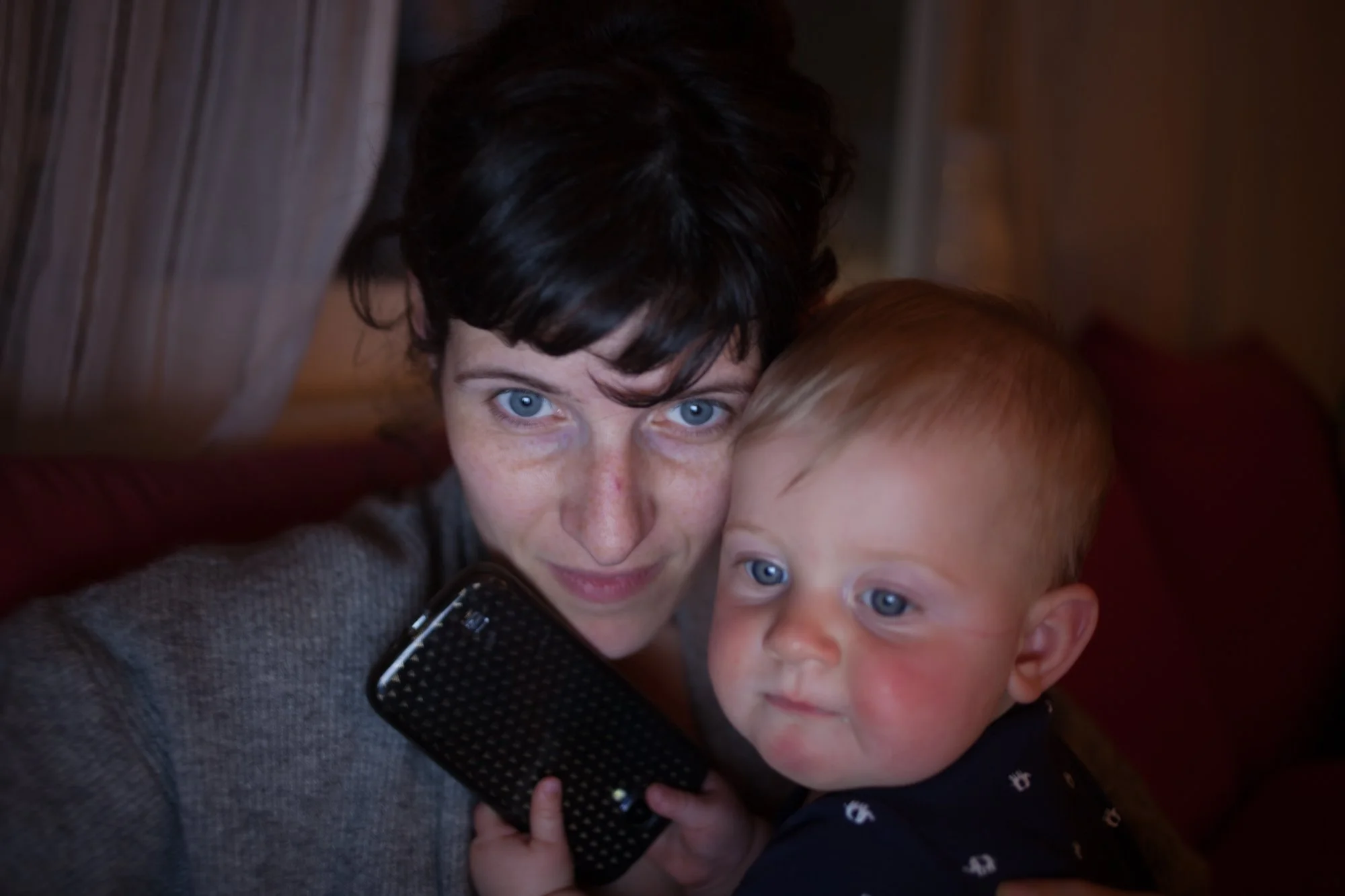 A woman with short dark hair and blue eyes holding a young blond-haired child with blue eyes, both focused on a smartphone.
