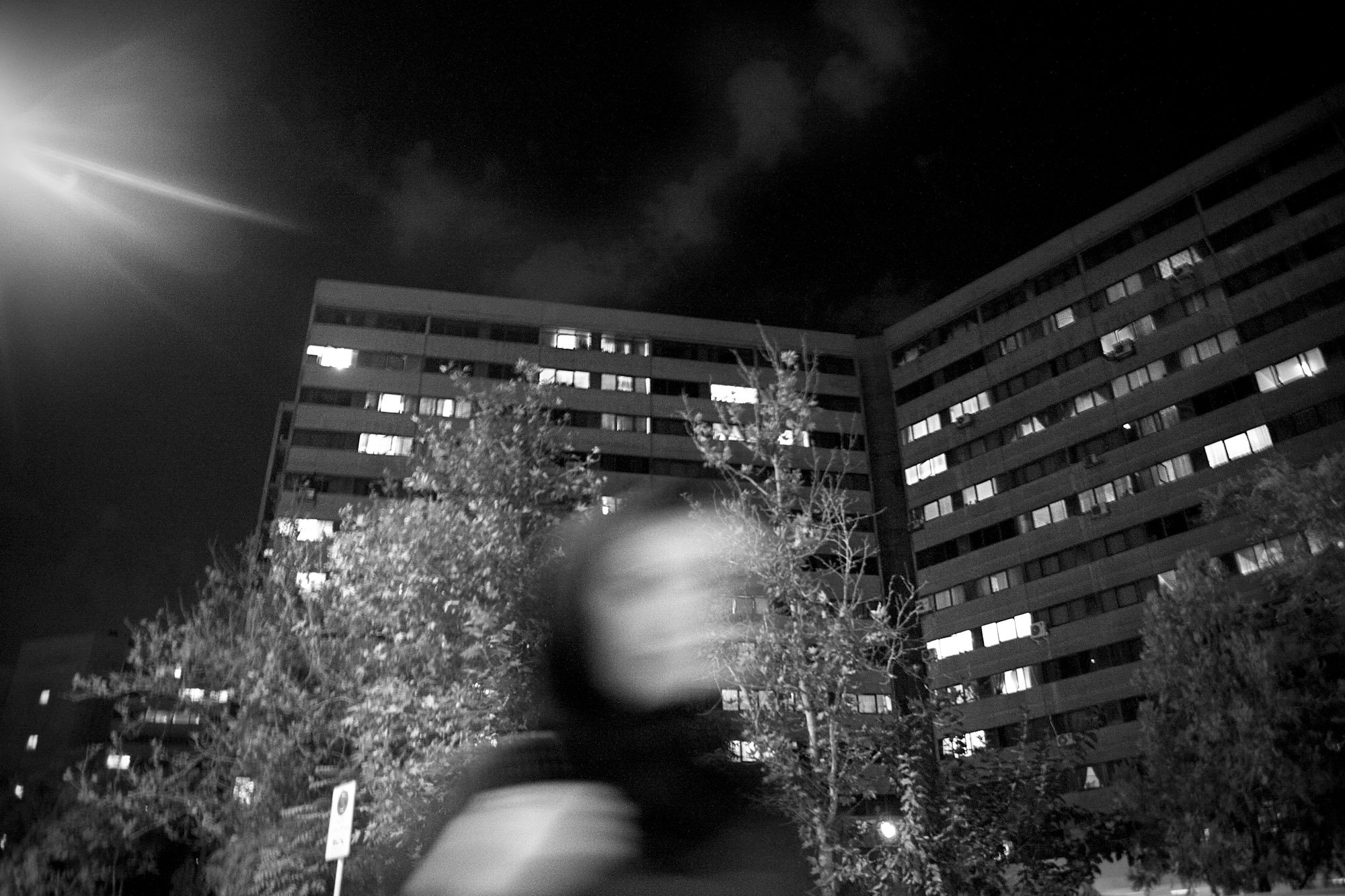 Black and white photo of a multi-story residential building at night, with windows illuminated, trees in the foreground, and a blurred figure in the lower part of the image.