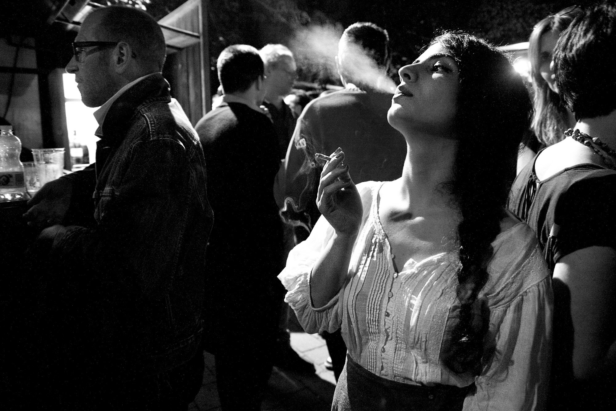 A black and white photo of a woman with long hair and a braid, holding a cigarette, standing among a group of people at a social gathering. The woman appears to be looking upward with a relaxed expression, while others around her are engaged in conve