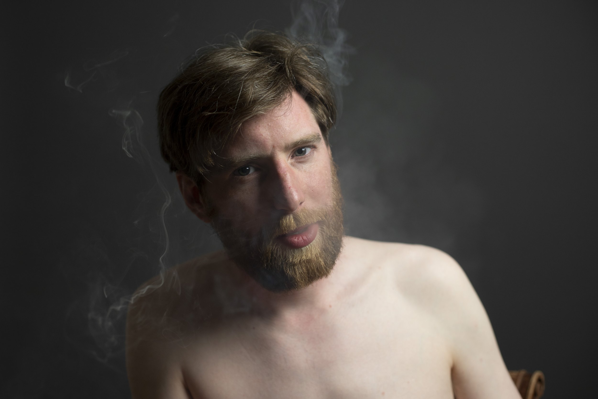 A shirtless man with brown hair and a beard, looking at the camera, with smoke around his head, against a dark background.