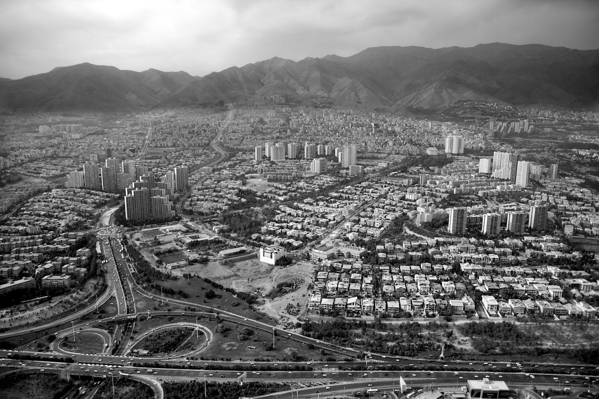Aerial view of a city with high-rise buildings, roads, and mountains in the background, in black and white.
