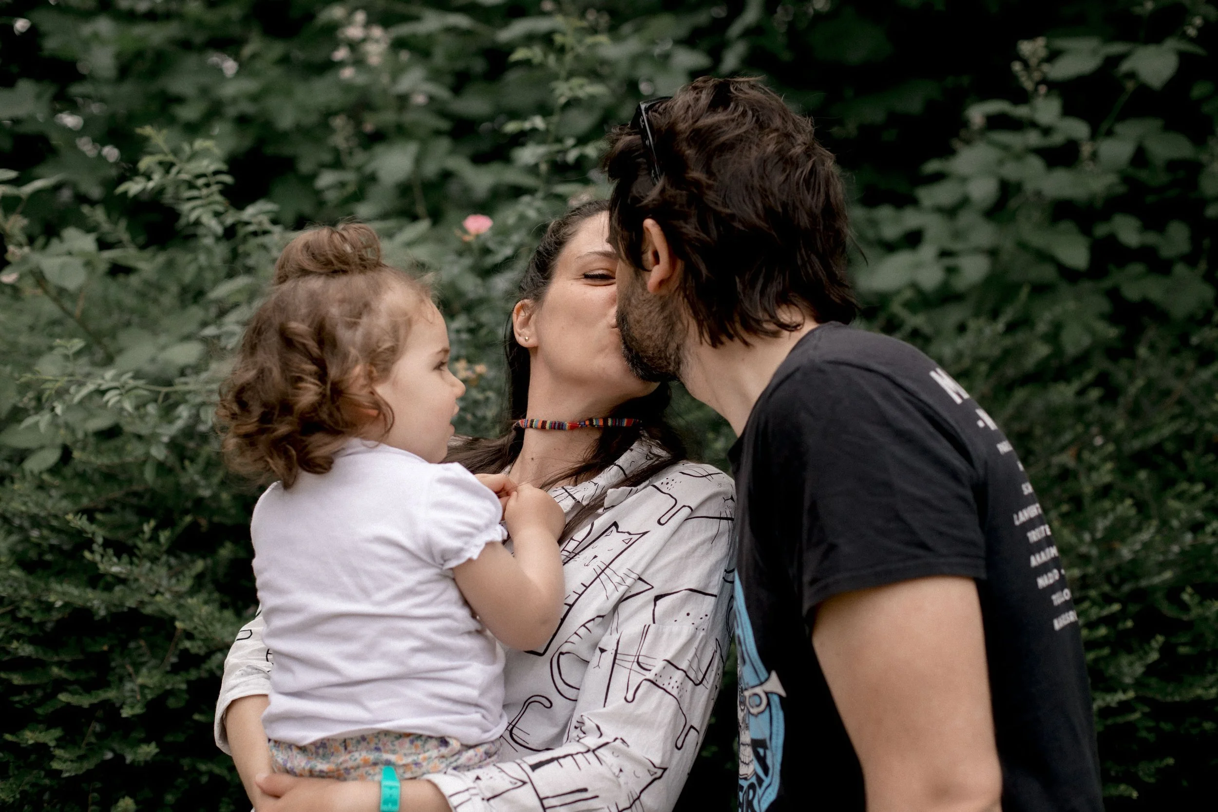A family outdoors with a woman holding a young girl, as the woman and a man kiss.