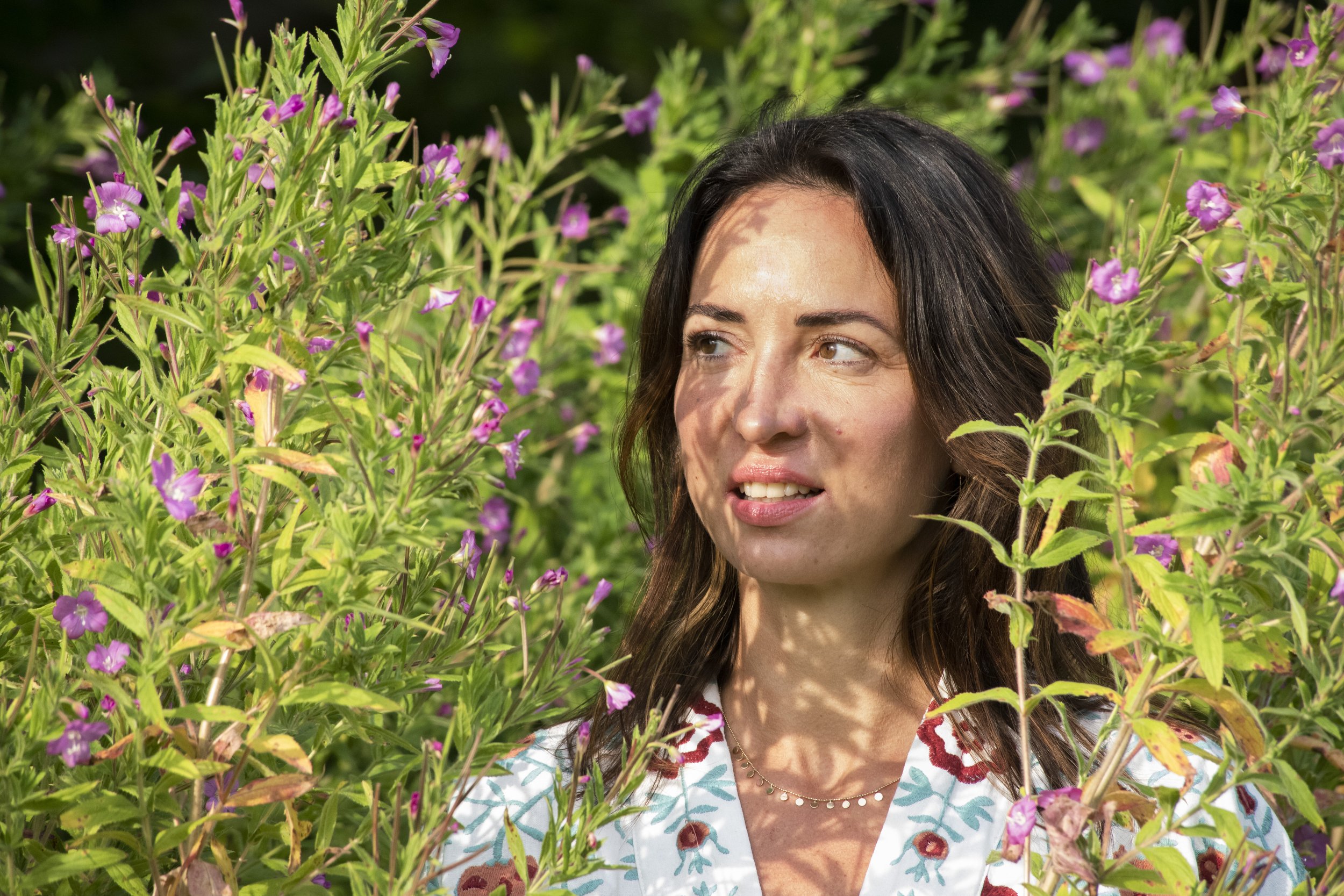 A woman with dark hair and a patterned blouse standing among green bushes with pink and purple flowers, sunlight and shadows on her face.