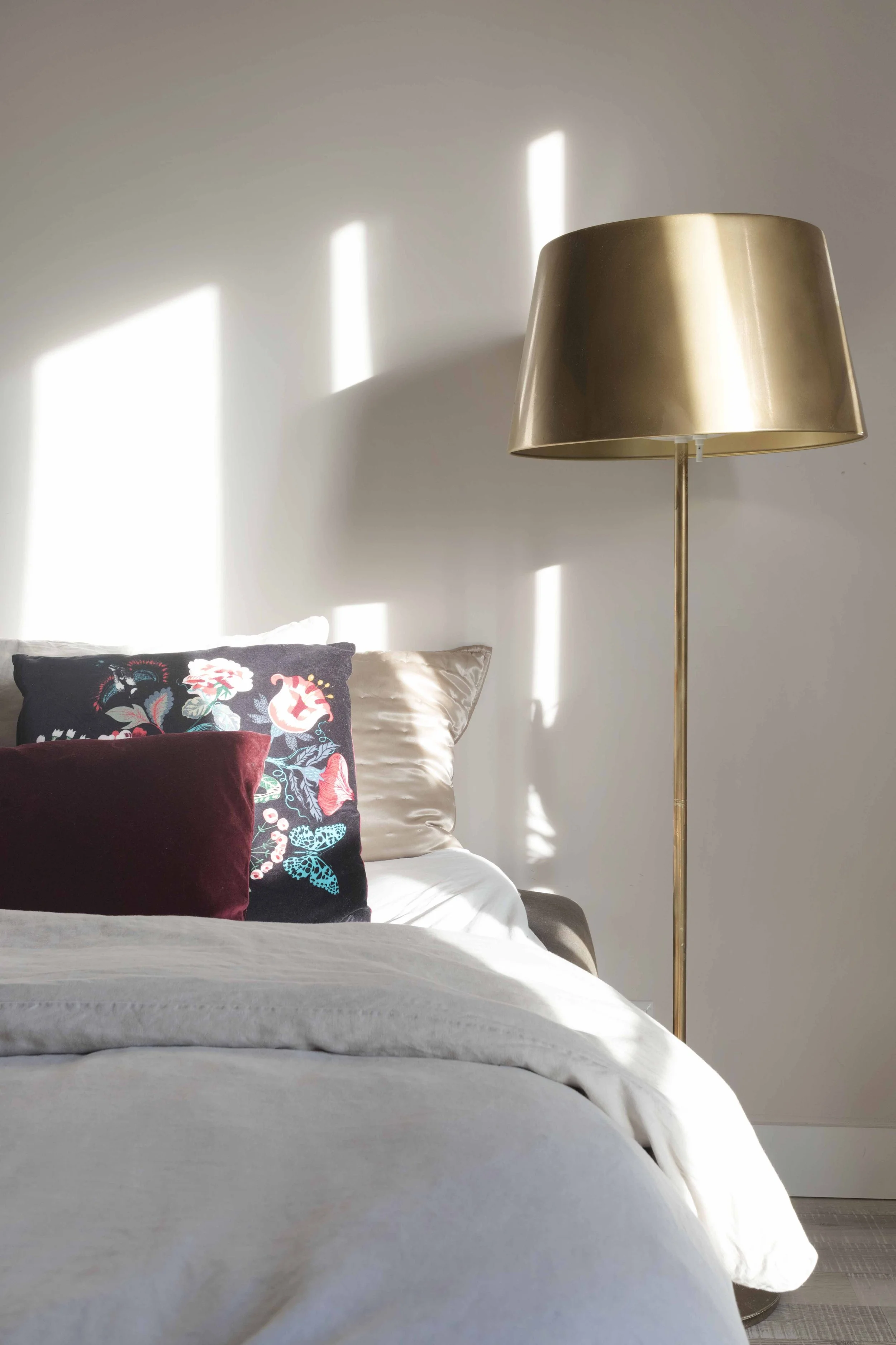 A bedroom with a bed, decorative pillows, and a brass floor lamp. Sunlight creates rectangular light patterns on the wall.
