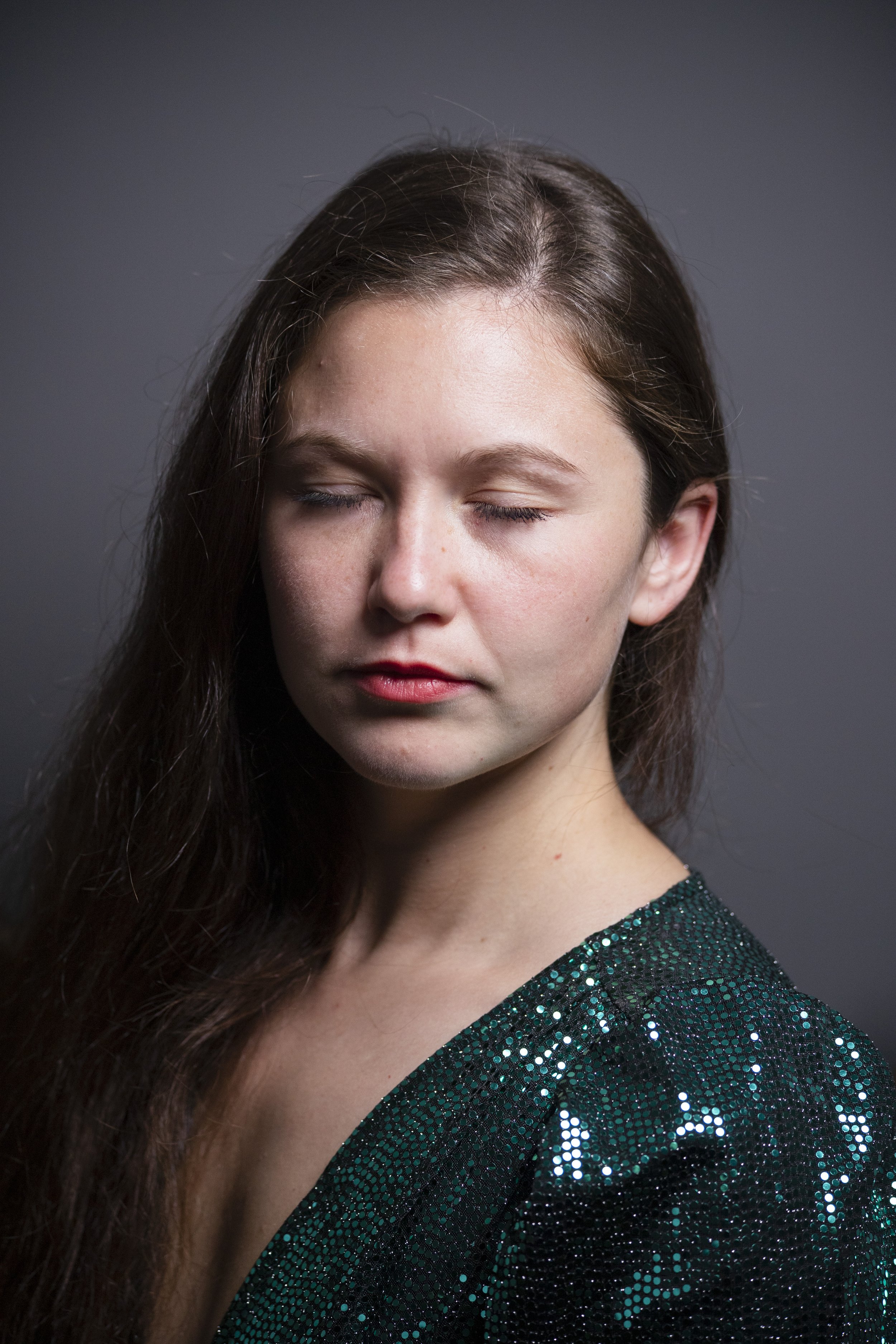 A young woman with long brown hair and closed eyes, wearing a shimmering green top, against a gray background.