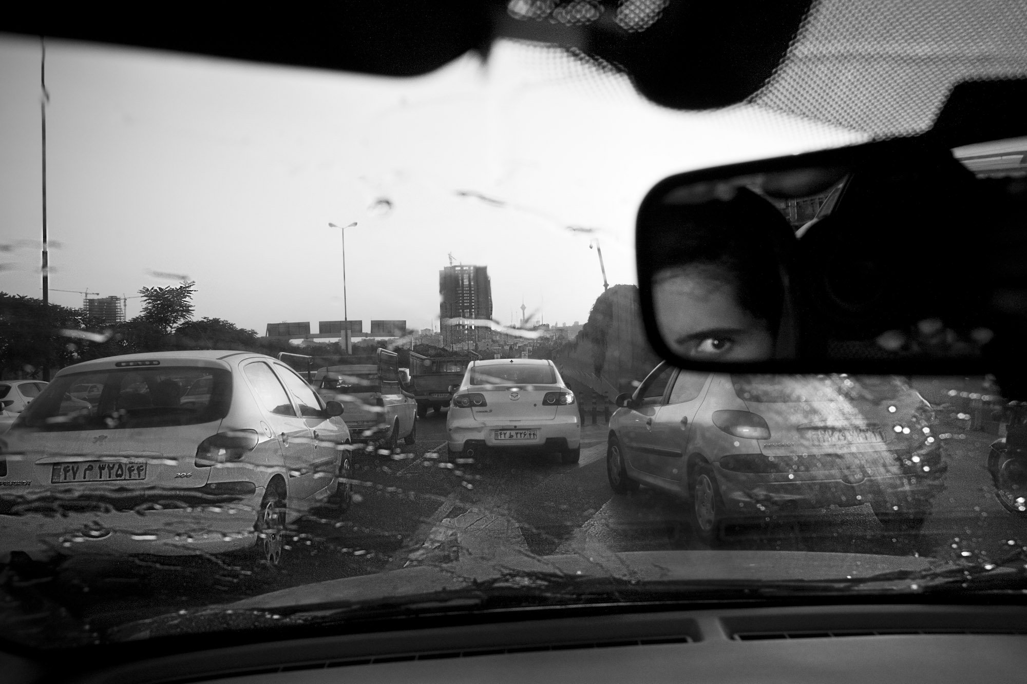 View through a rain-covered windshield showing a parking lot with multiple cars and a cityscape in the background. The photo is in black and white, and a person's eye is visible in the rearview mirror reflection.